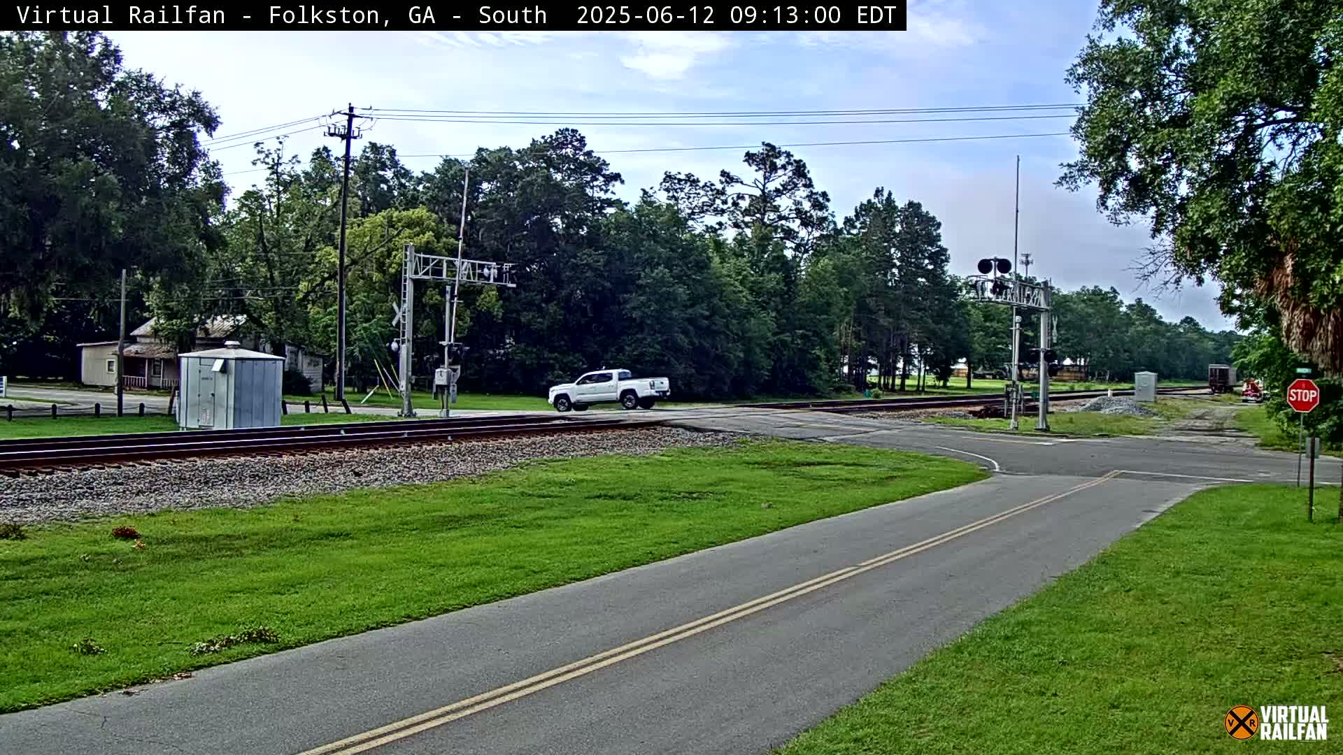 A white pickup truck is stopped at a railroad crossing on a partly cloudy day, with lush green grass and trees surrounding the area.