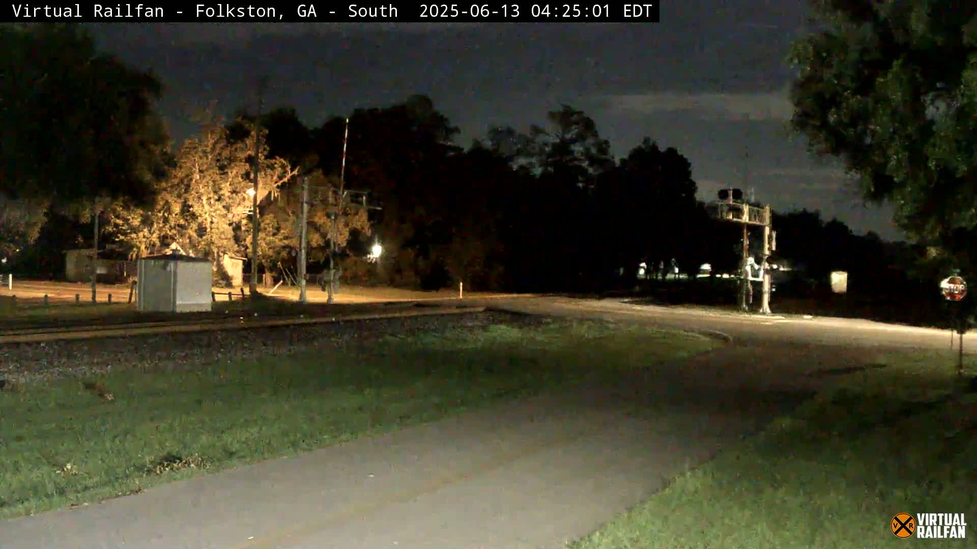 A nighttime view of a railroad crossing with a small building and trees under a dark sky.