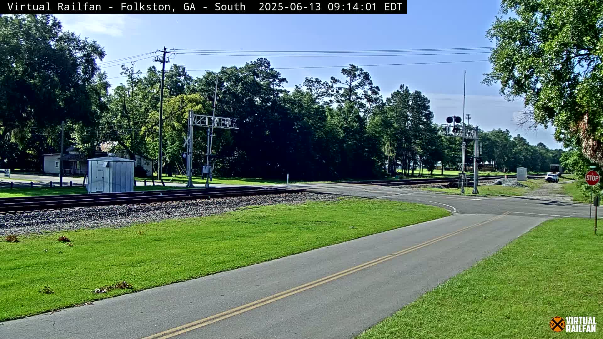 A mostly sunny day shows a railroad crossing with a road intersecting, surrounded by green grass and trees.