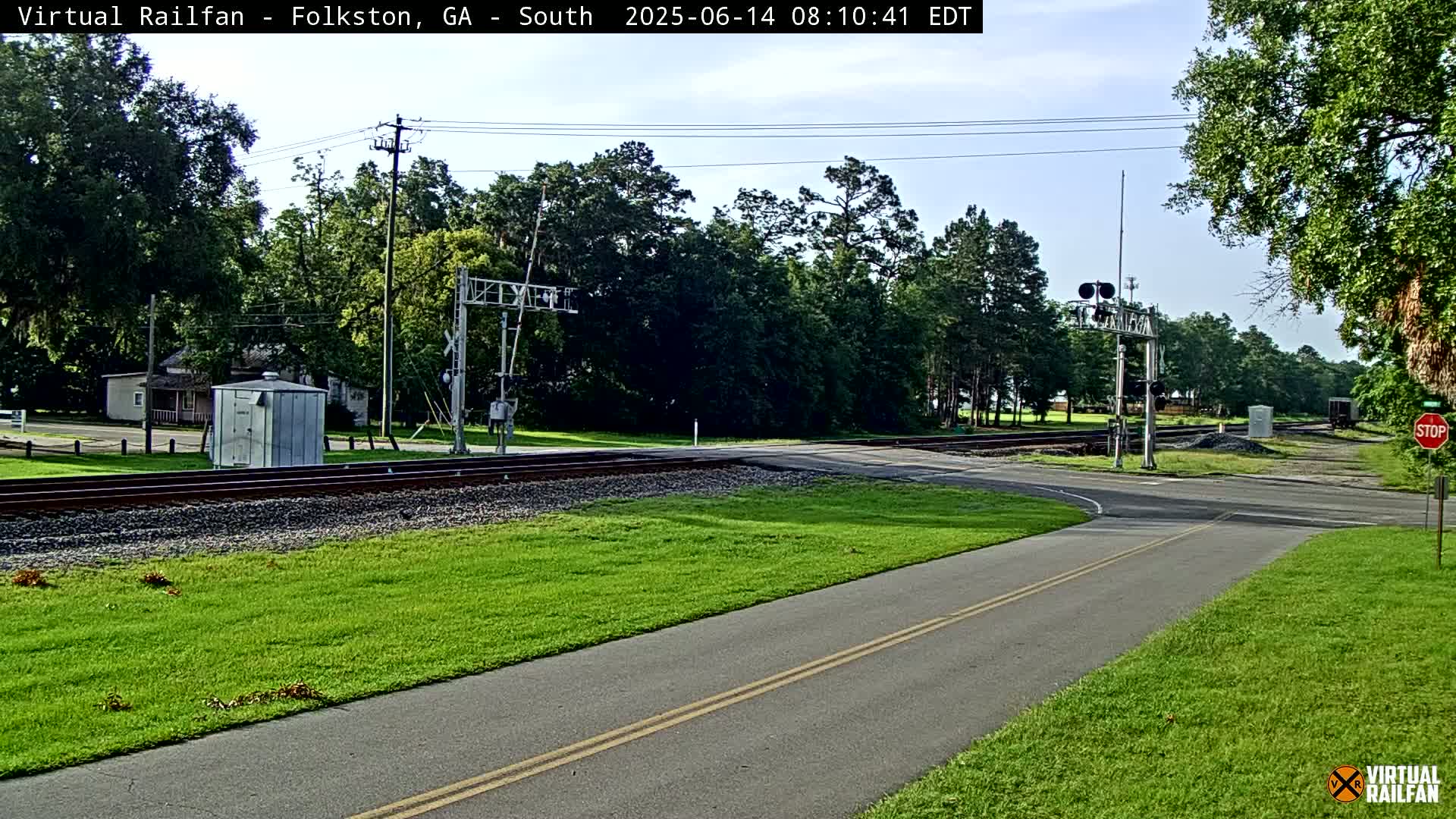 A paved path intersects with a railroad crossing under a mostly clear sky, surrounded by green grass and trees.