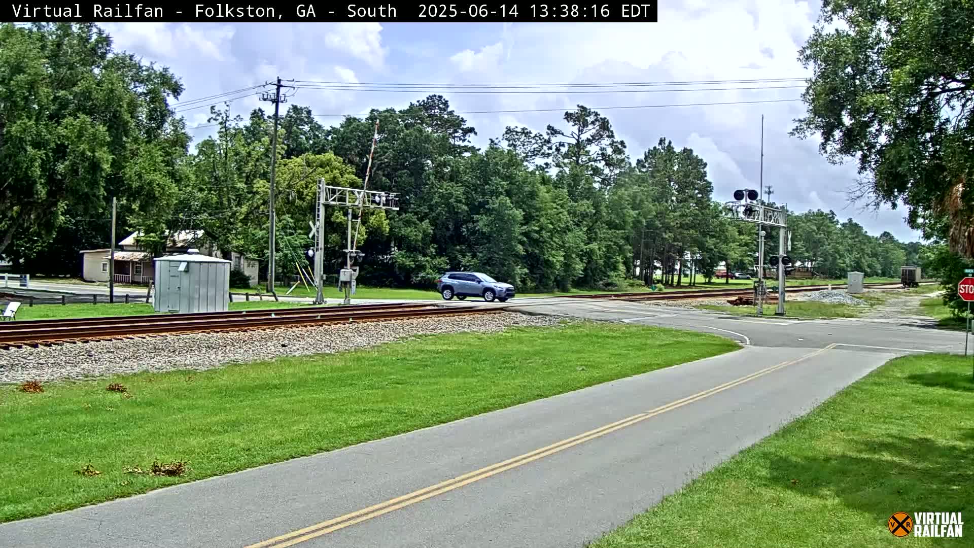 A gray SUV is crossing a railroad track at a road intersection, surrounded by green grass and trees under partly cloudy skies.