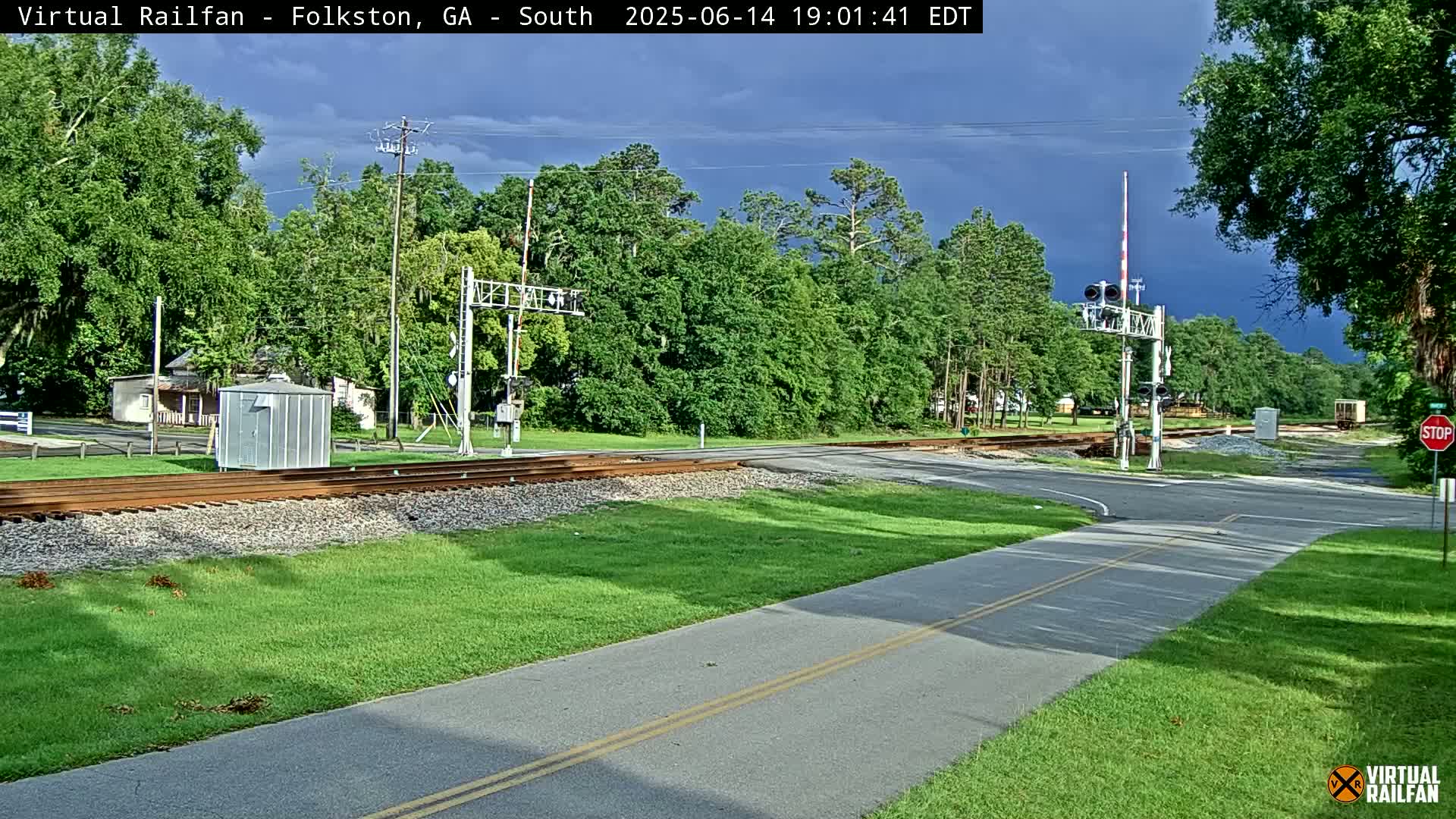 A gray SUV is crossing a railroad track at a road intersection, surrounded by green grass and trees under partly cloudy skies.