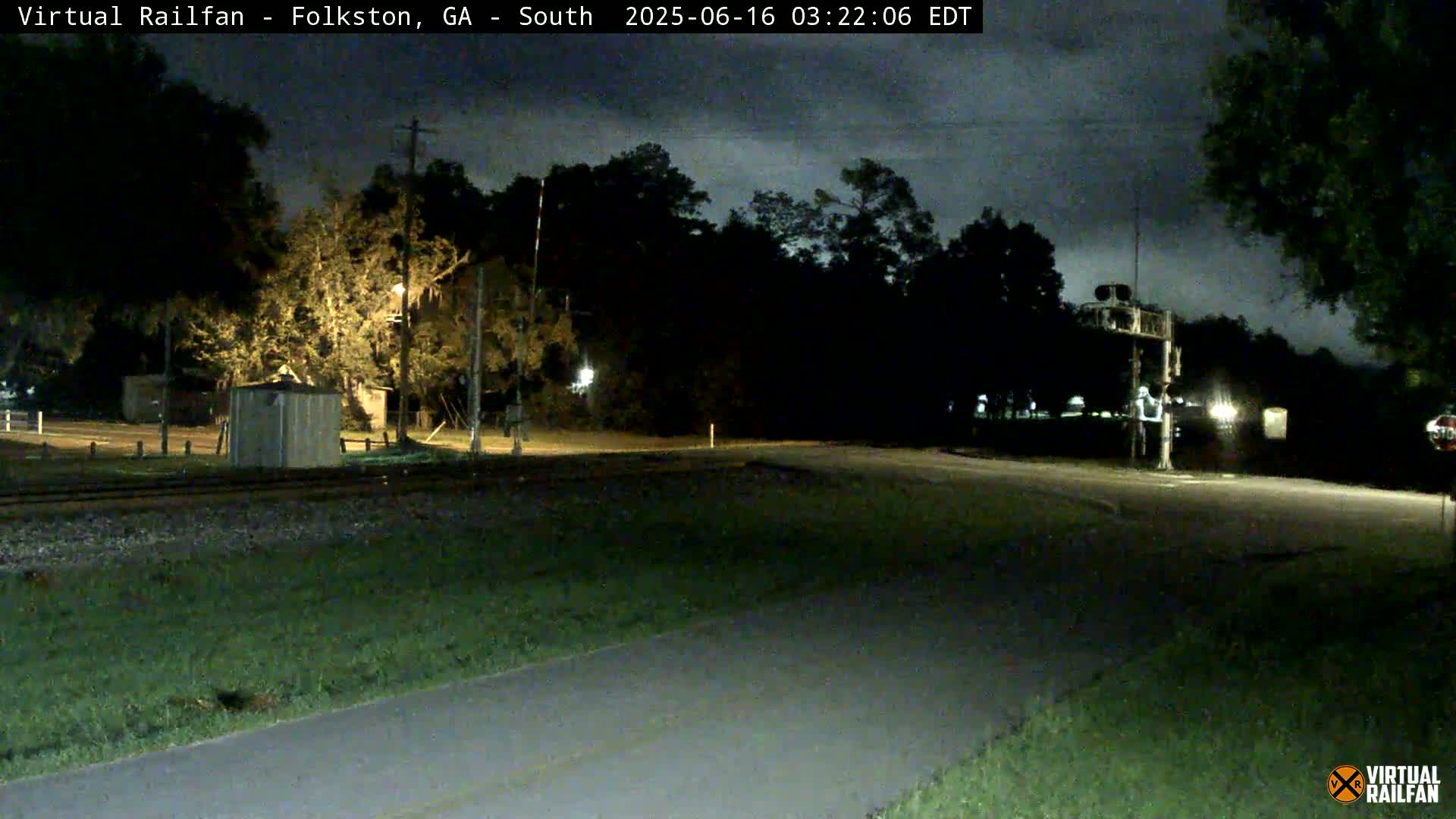 A nighttime view of a railroad crossing with a road intersecting, surrounded by trees under a partly cloudy sky.