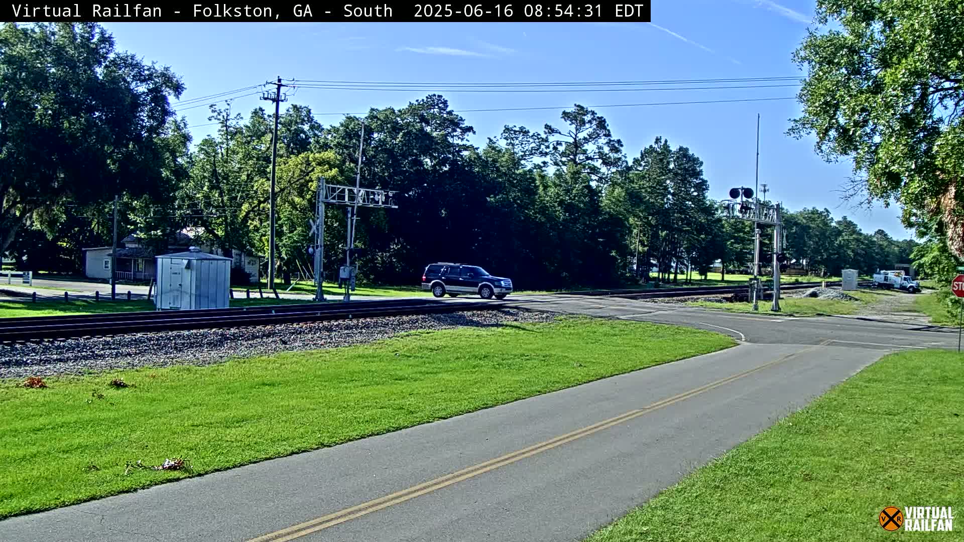 A dark-colored SUV drives across a railroad crossing on a sunny day, passing through a rural intersection with lush green grass and trees.