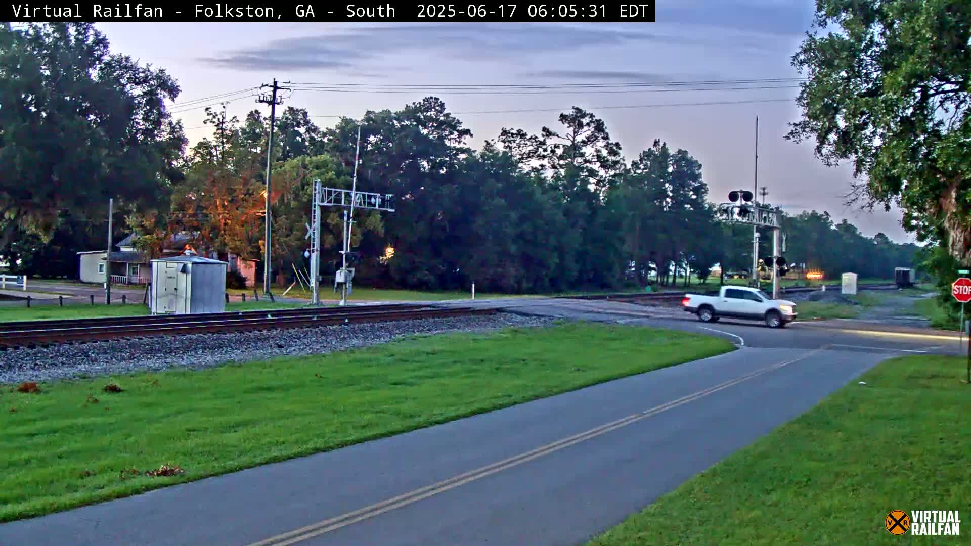 A white pickup truck turns onto a road that crosses a set of railroad tracks under a twilight sky.