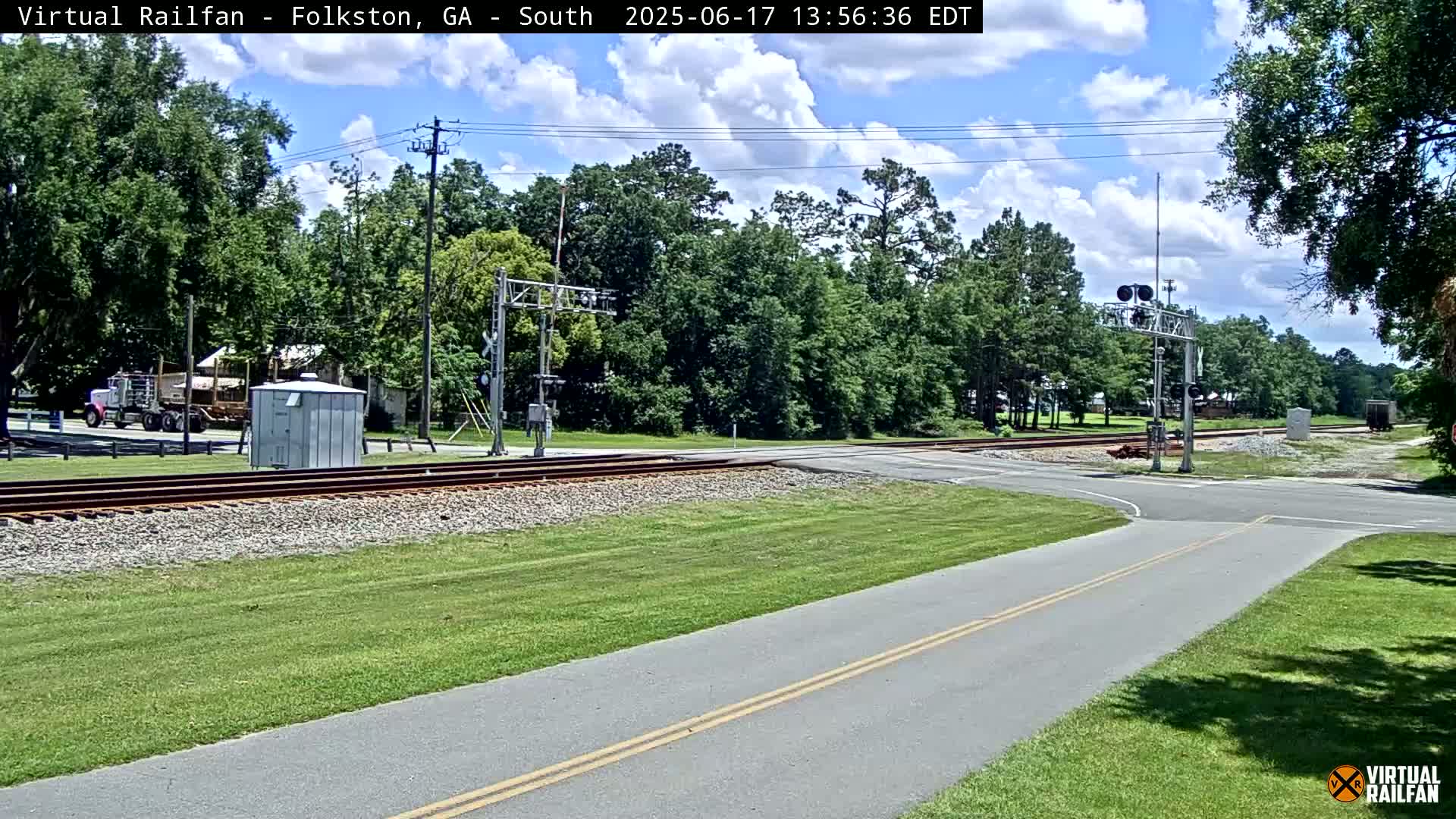 A paved path intersects a road crossing railroad tracks under a partly cloudy sky, with green grass and trees surrounding the area.