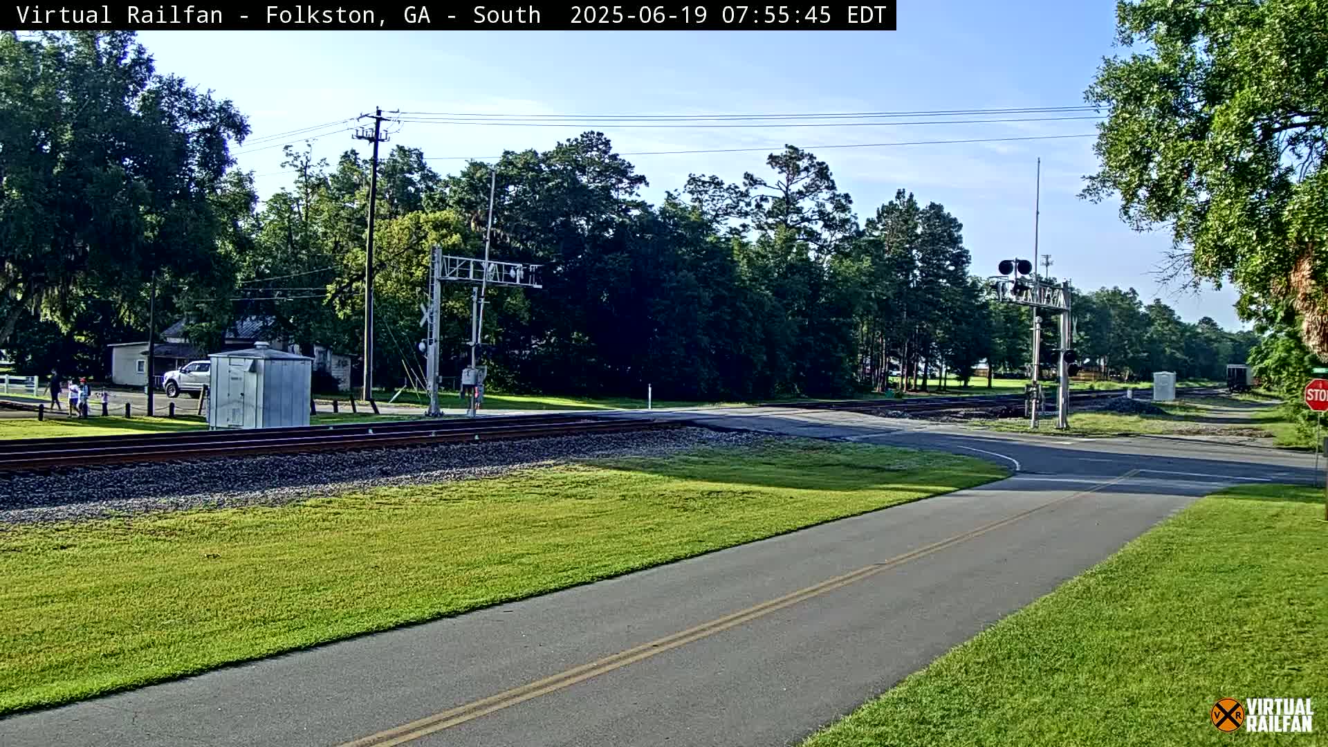 A paved road intersects with a railroad crossing under a mostly sunny sky, surrounded by green grass and trees.