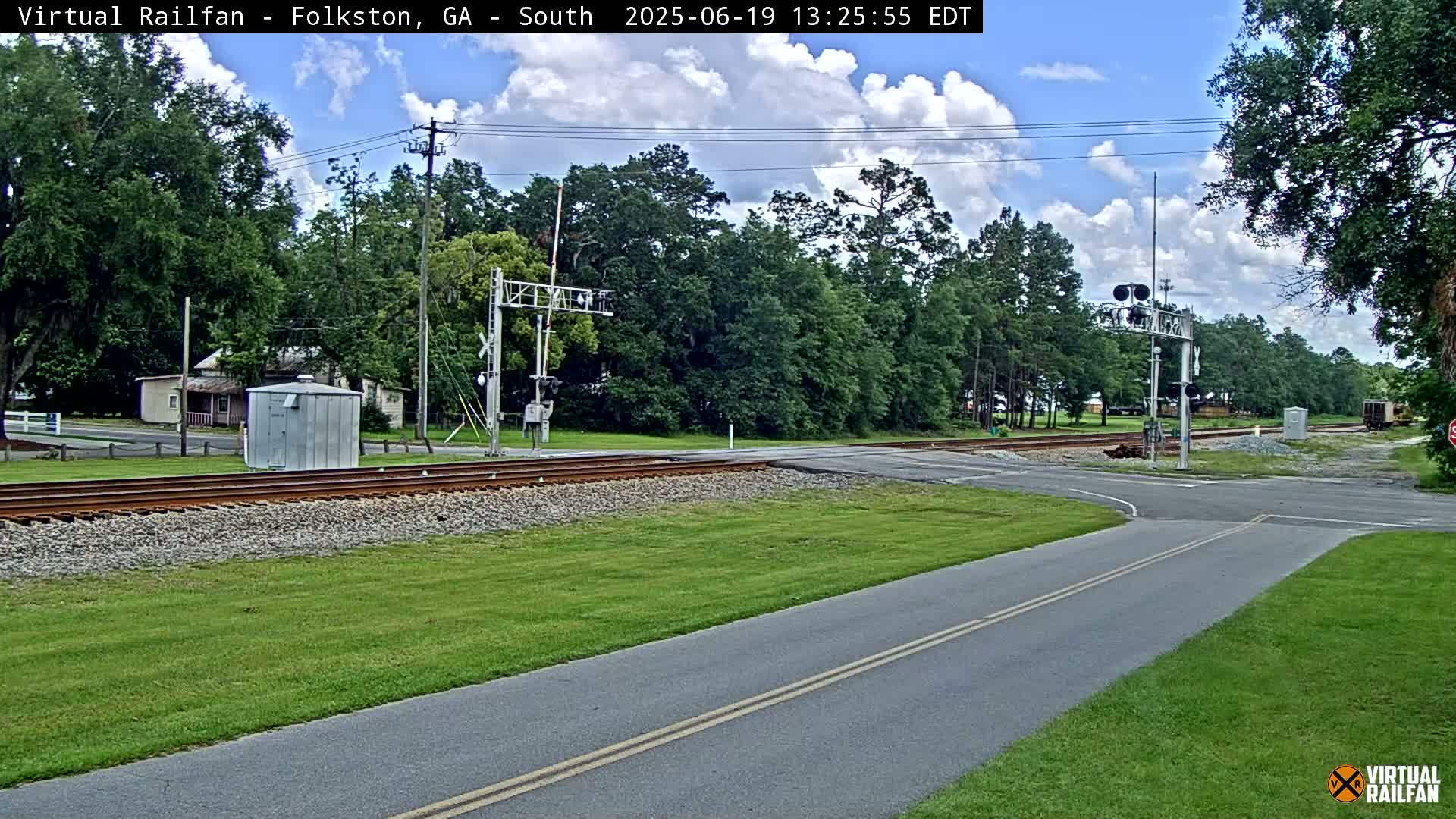 A railroad crossing with a road intersecting the tracks under partly cloudy skies, surrounded by green grass and trees.