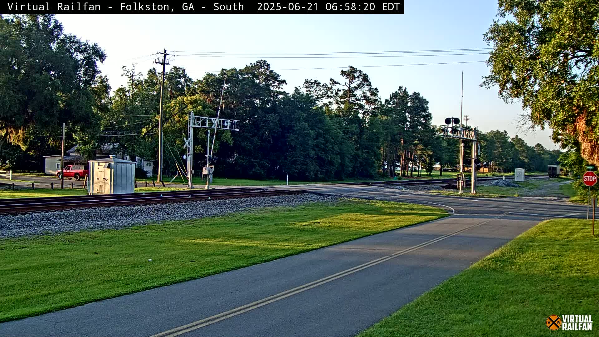 A paved path and road intersect at a railroad crossing under a partly sunny sky, surrounded by green grass and trees.