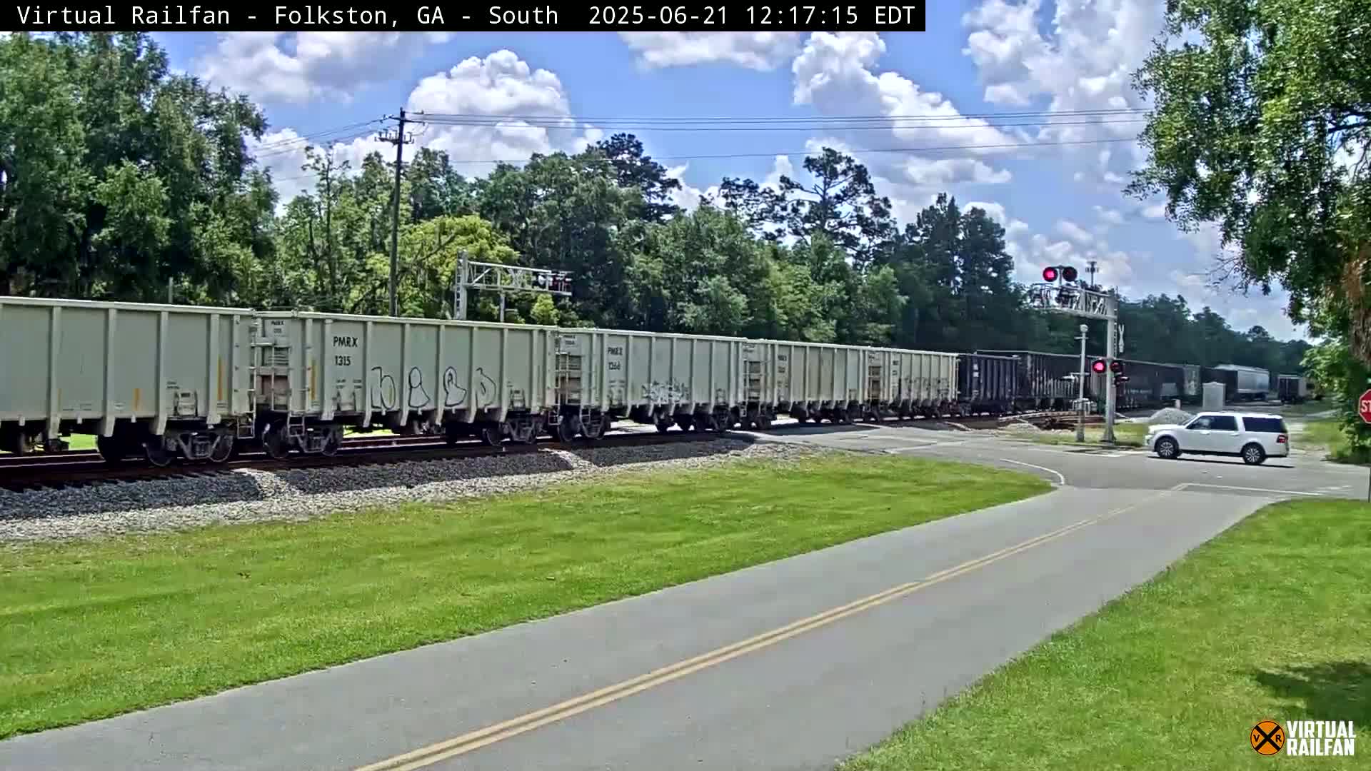 A long freight train passes a road crossing under a partly cloudy sky, with a white SUV waiting at the intersection.