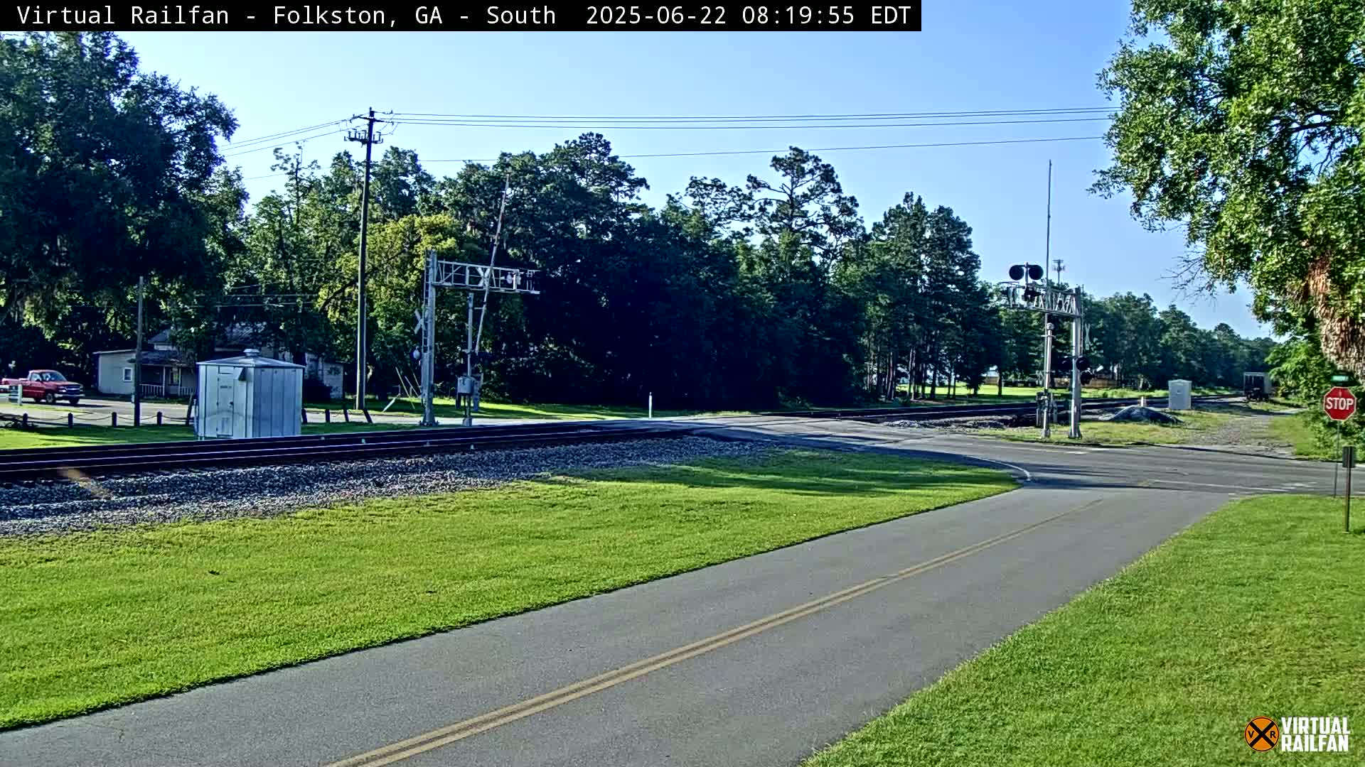 A paved path and road intersect at a railroad crossing under a clear sky, with lush green grass and trees lining the area.