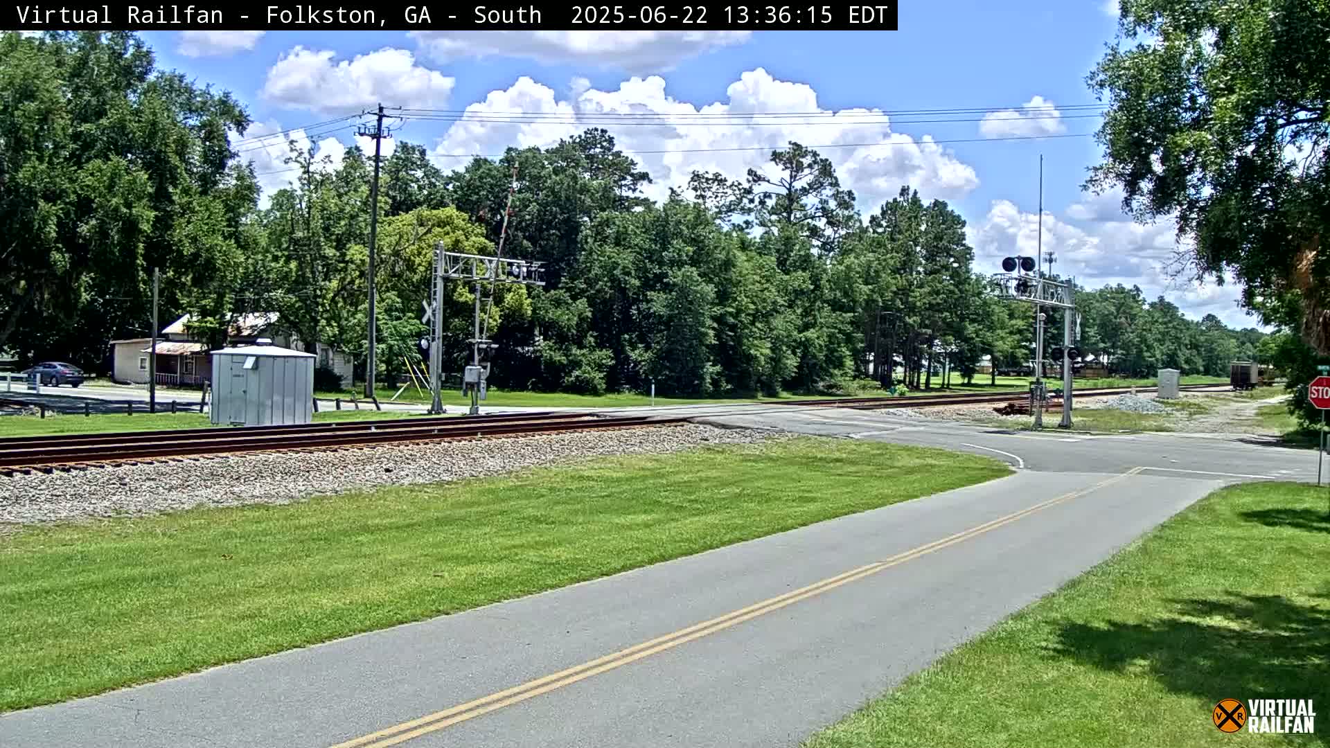 A paved road with double yellow lines intersects a railroad crossing under a partly cloudy sky, surrounded by green grass and trees.