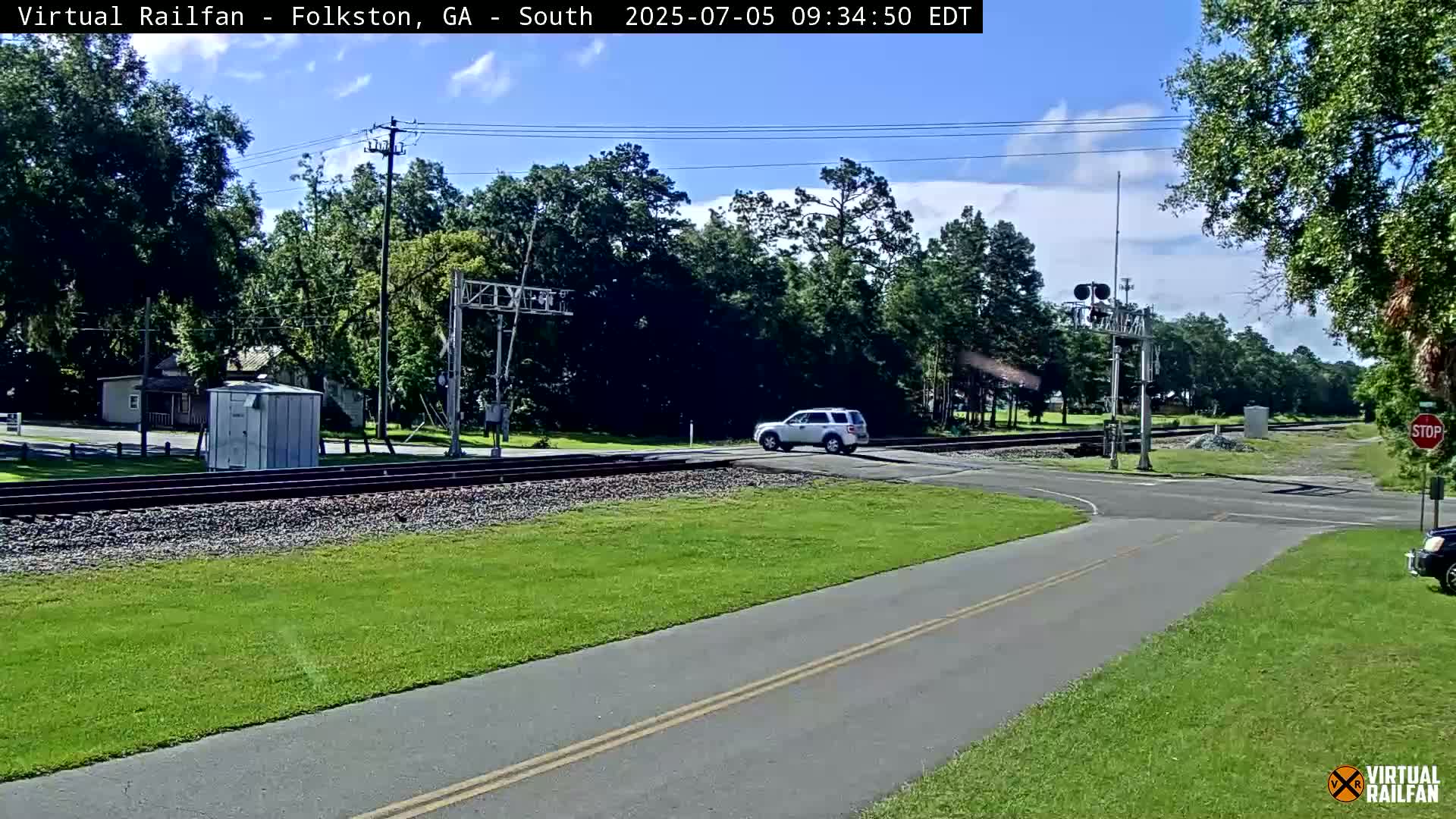 A partly cloudy day shows a white SUV crossing a railroad track at a road intersection, with lush green grass and trees surrounding the area.