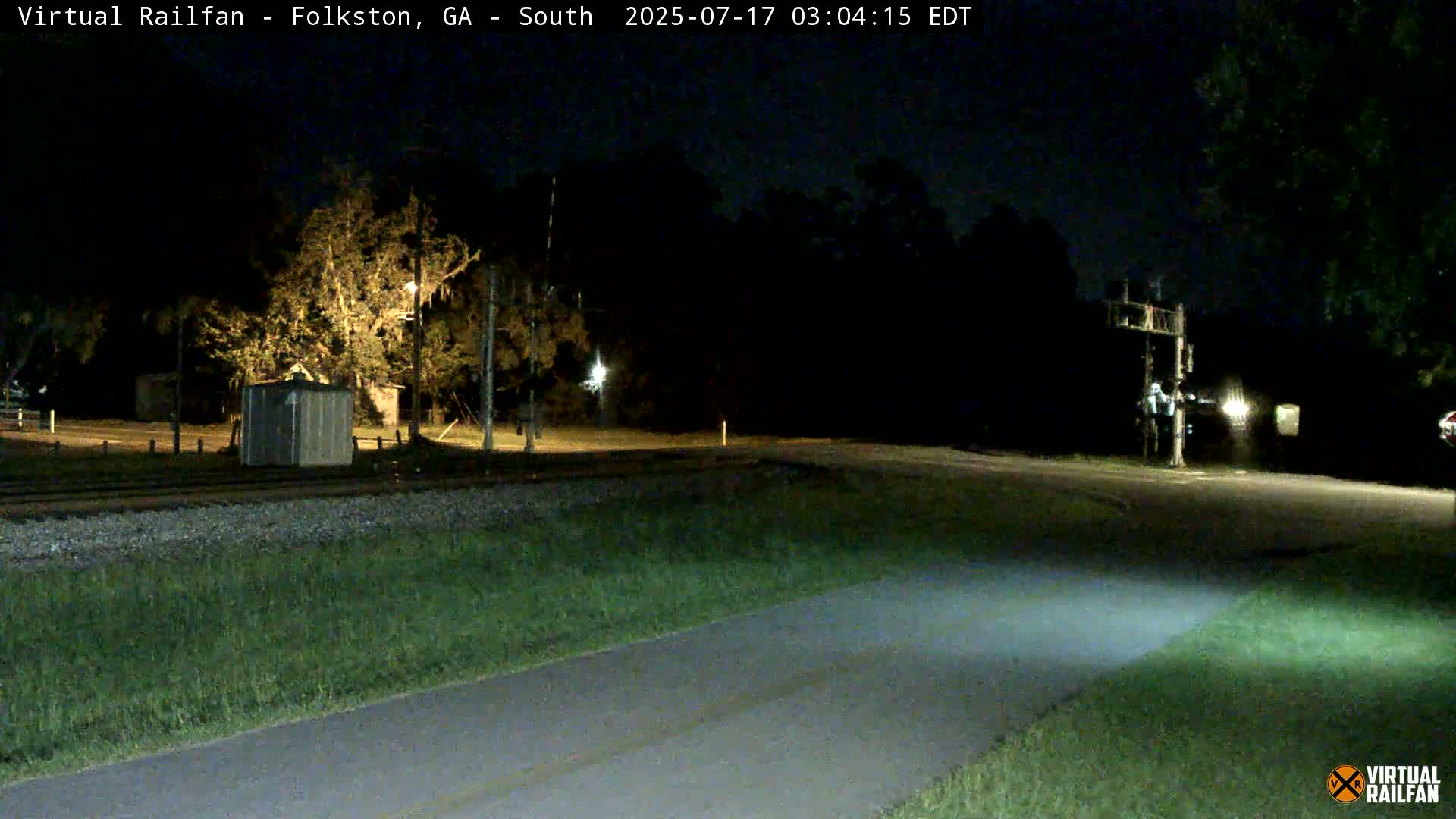 A paved path stretches toward a nighttime intersection near railroad tracks and trees under a clear, dark sky.