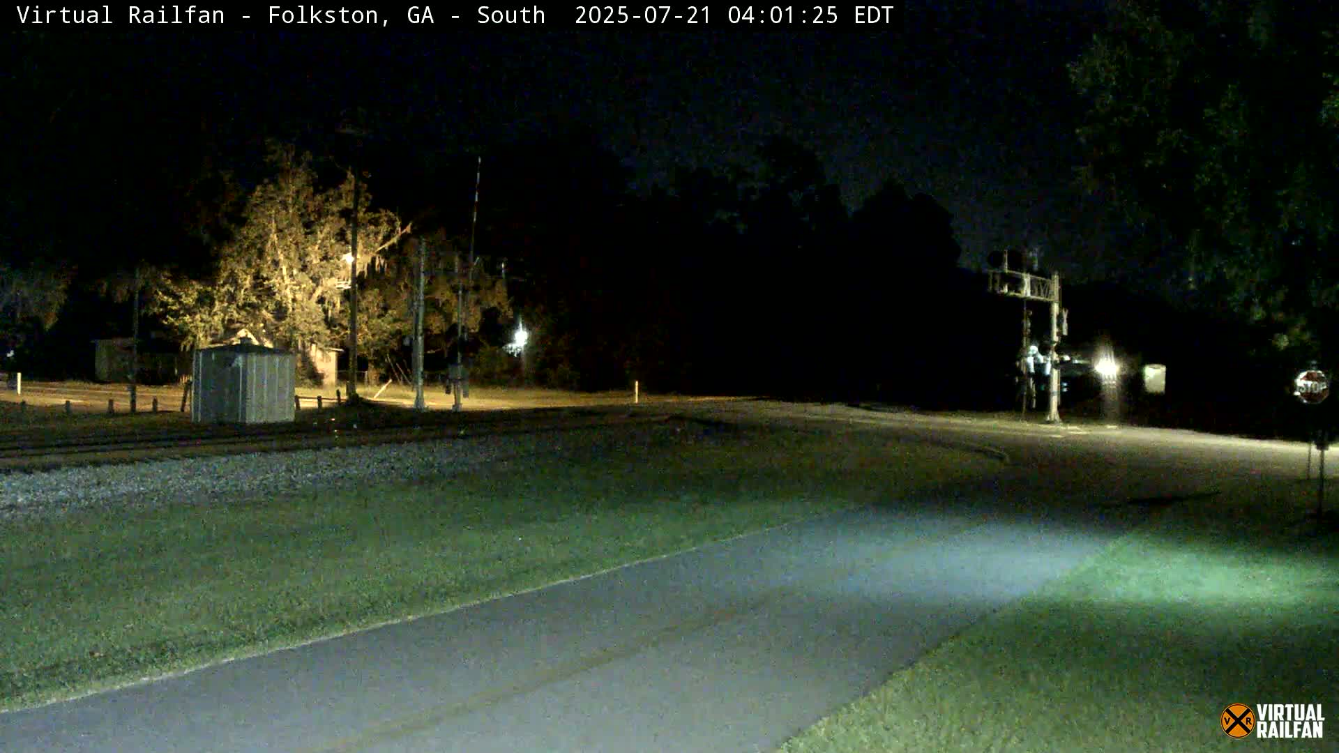 A nighttime view of a railroad crossing with a road and a paved path, under dark, clear skies.