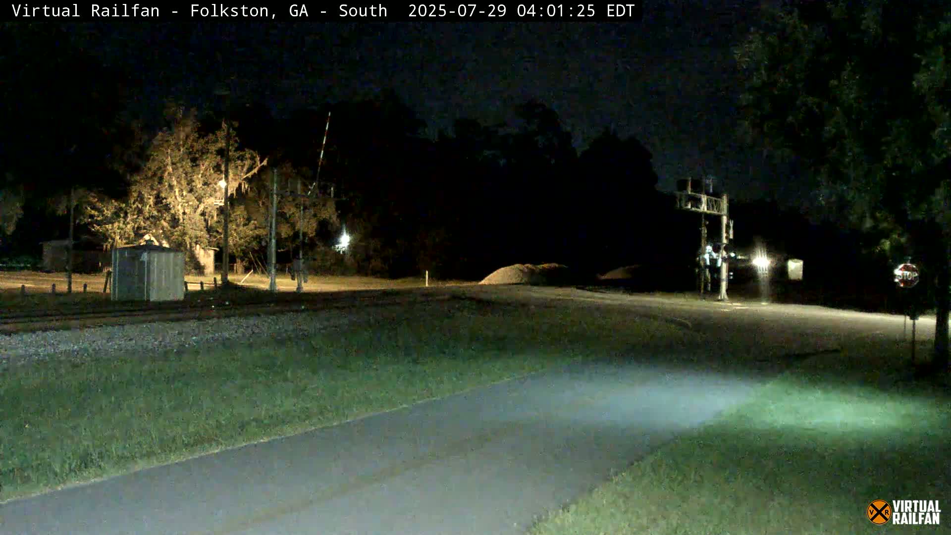 A nighttime view of a road intersecting with railroad tracks, under a clear sky, showing a small shed and trees near the tracks.