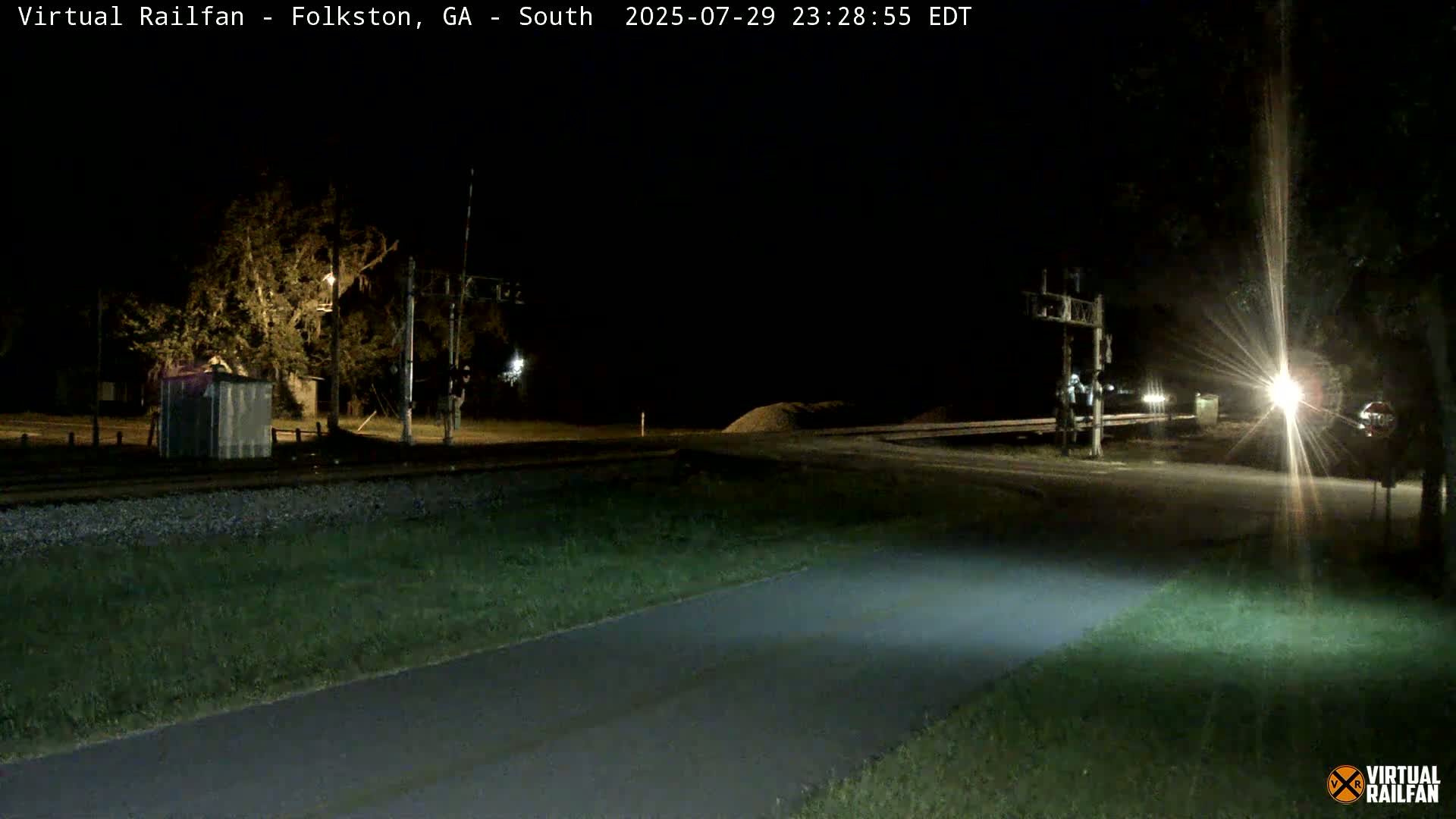 A nighttime view of a road intersecting with railroad tracks, under a clear sky, showing a small shed and trees near the tracks.