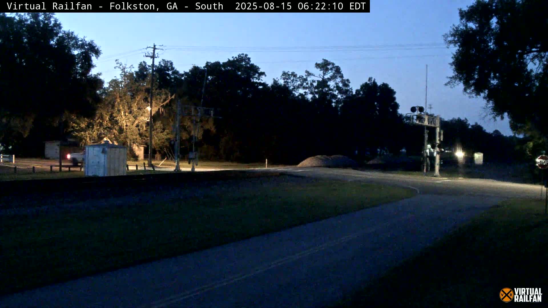 A nighttime view of a road intersecting with railroad tracks, under a clear sky, showing a small shed and trees near the tracks.