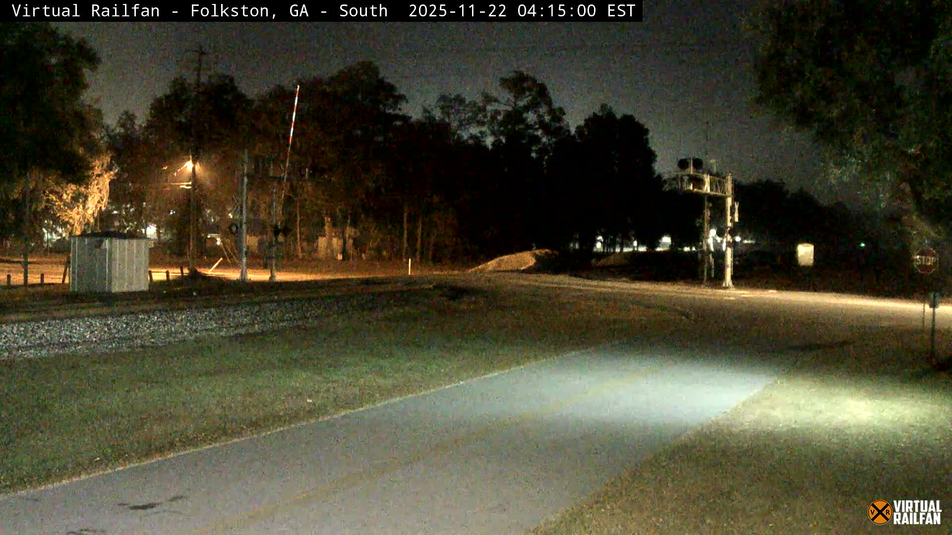 A clear night view shows a railroad crossing with tracks, signals, and a streetlamp illuminating a road and grassy area, surrounded by dark trees.