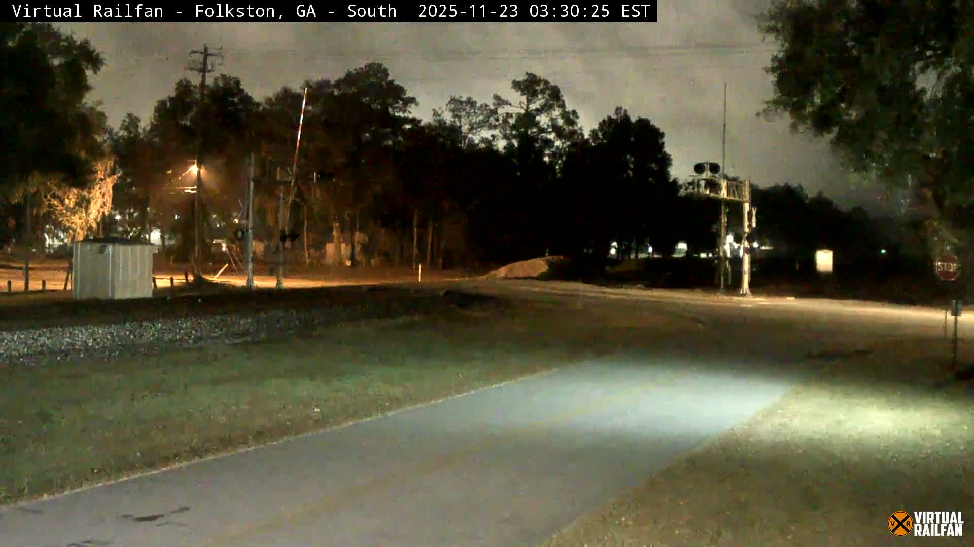 A nighttime view shows a railroad crossing with a raised gate arm and signal structure, flanked by a paved path, grassy areas, a utility building, and trees, all under a cloudy sky.