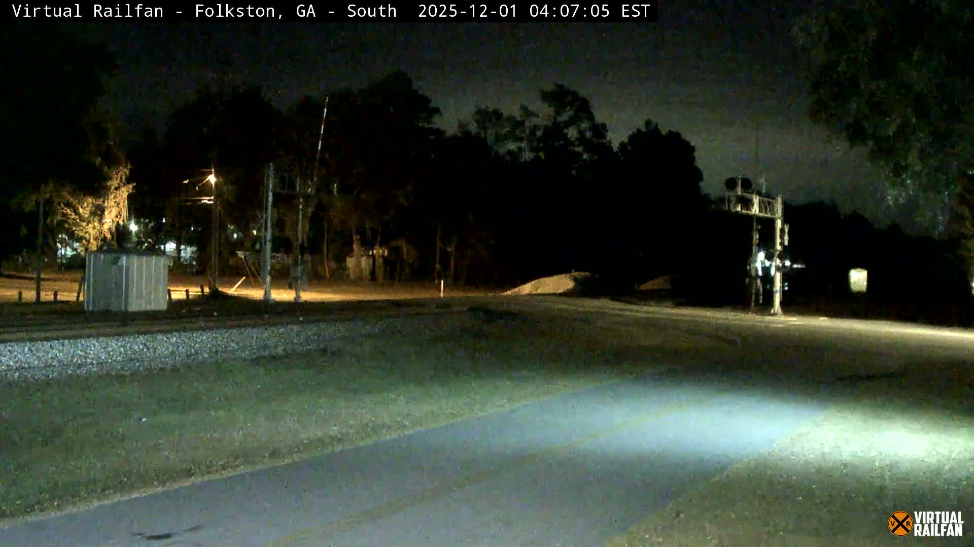 A nighttime outdoor scene features railroad tracks adjacent to a paved road, a small utility building on the left, and a railroad crossing signal on the right, all partially illuminated by artificial lights under a clear sky.