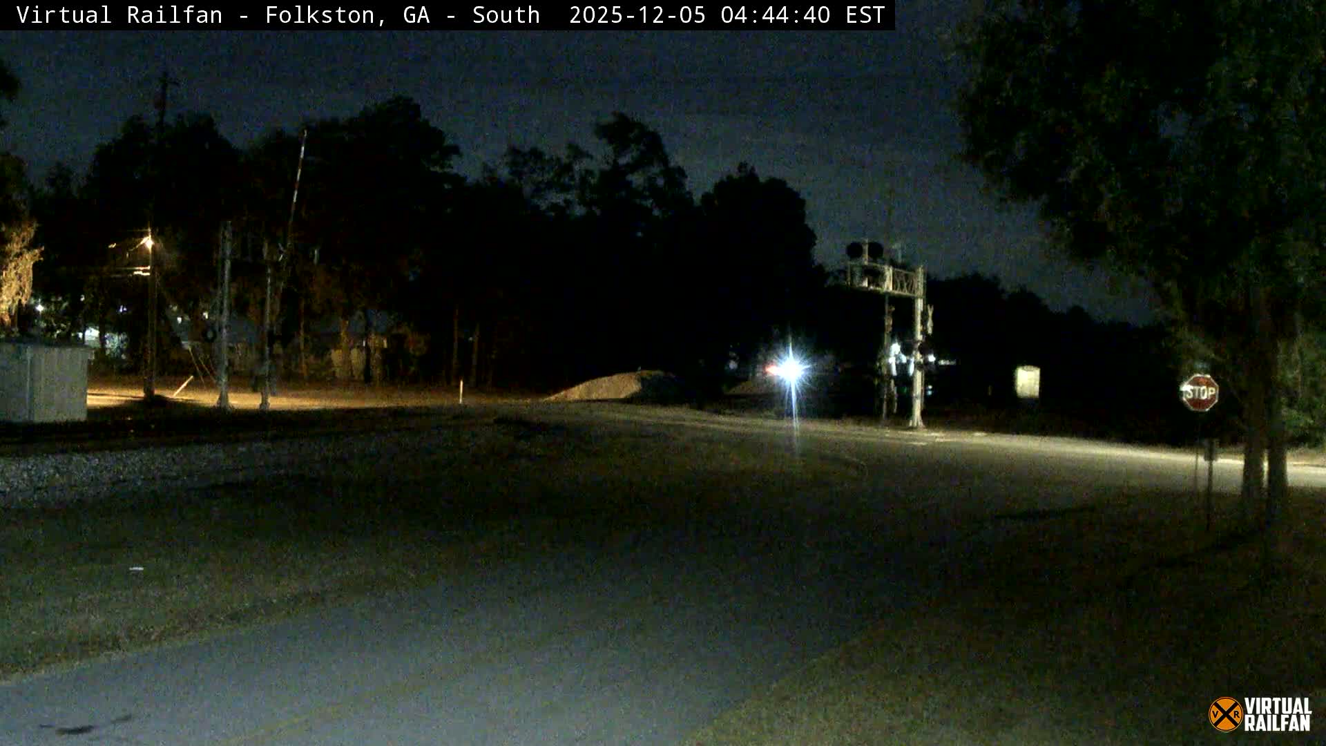 A dark night scene at a railroad crossing shows trees, a stop sign, and a bright light in the distance under clear skies.
