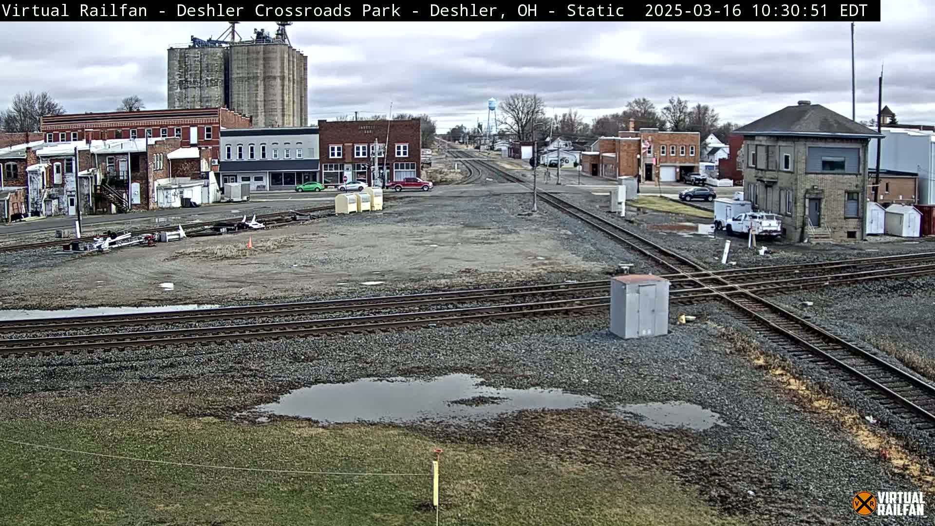 A view of a railroad crossing in a small town under overcast skies, with several buildings and a few cars visible.