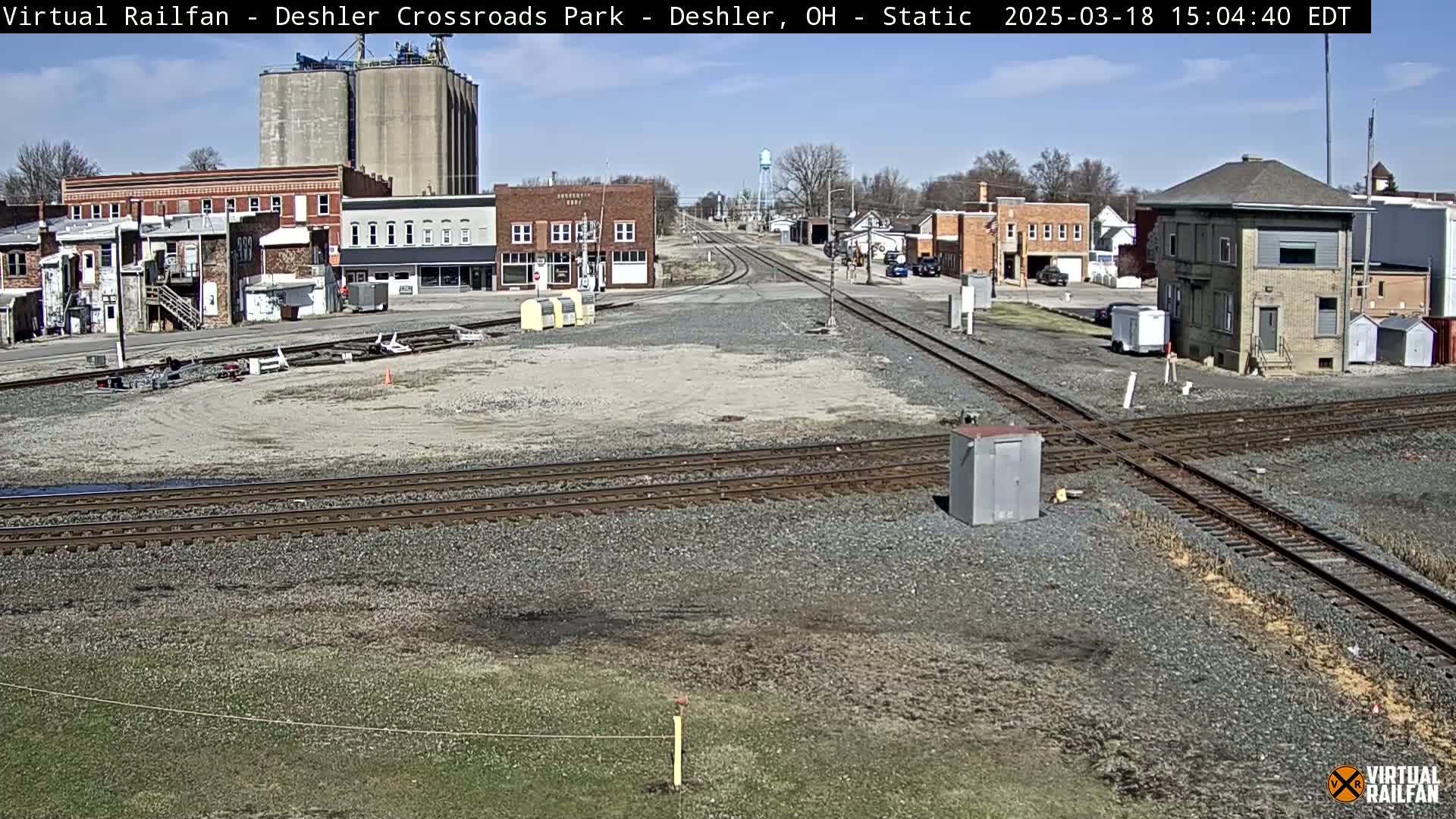 A sunny day reveals a railroad crossing in a small town, with several buildings, grain silos, and train tracks visible.