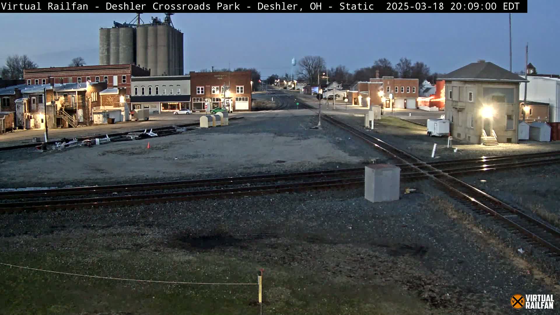 A twilight view of a railroad crossing in a small town, with buildings, grain silos, and train tracks visible under a clear, dark sky.