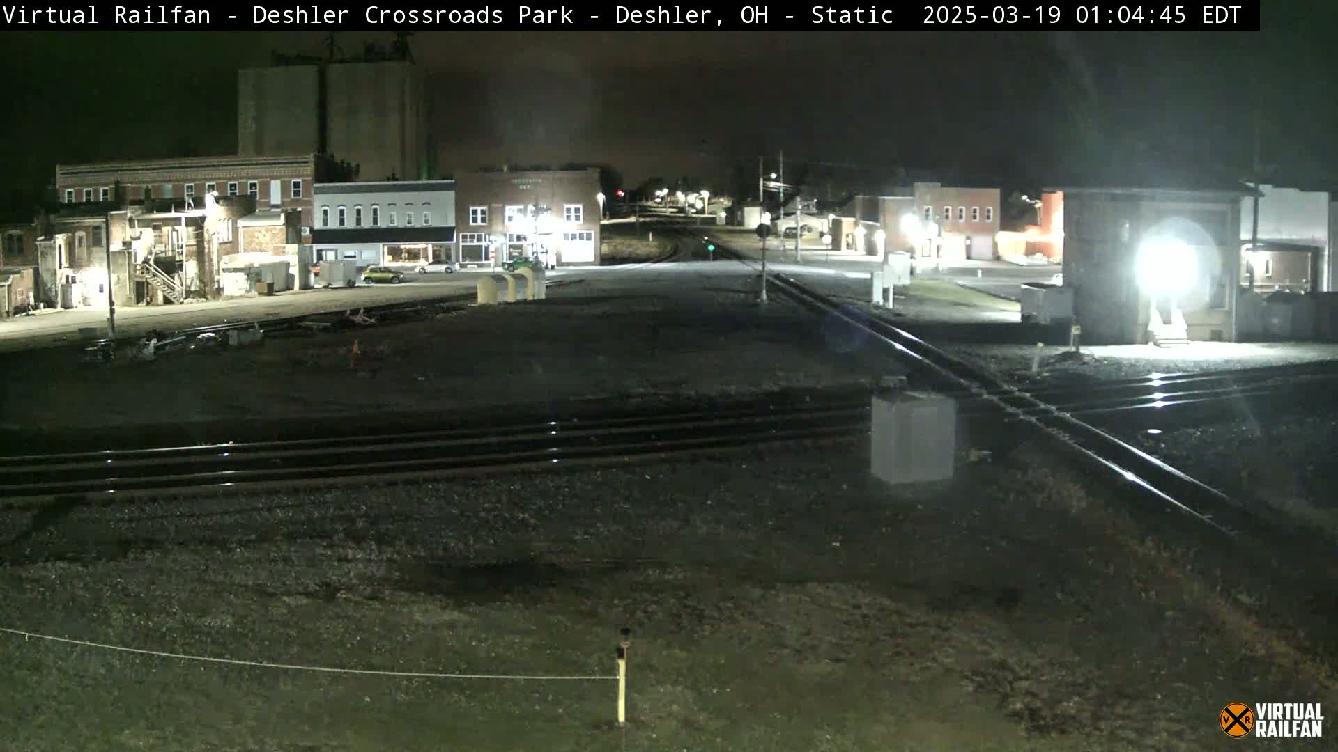 At night, a deserted train crossing in a small town features railroad tracks intersecting a road, with a few buildings visible under dim lighting.