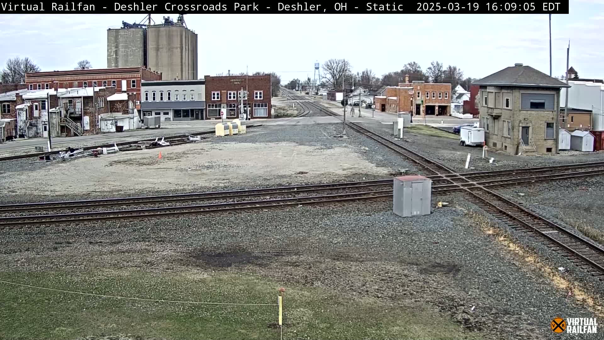 A mostly clear day view of a railroad crossing in a small town, featuring intersecting train tracks, several buildings, and a small, light-grey, square structure.