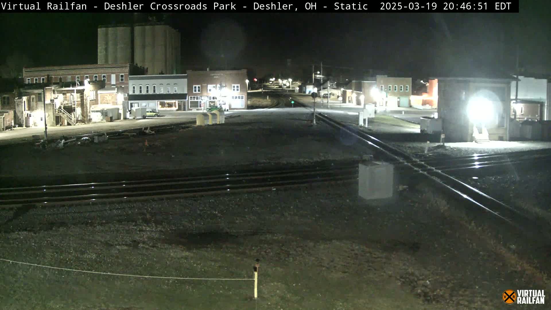 A nighttime view of a railroad crossing in a small town, with several buildings visible under dark skies.