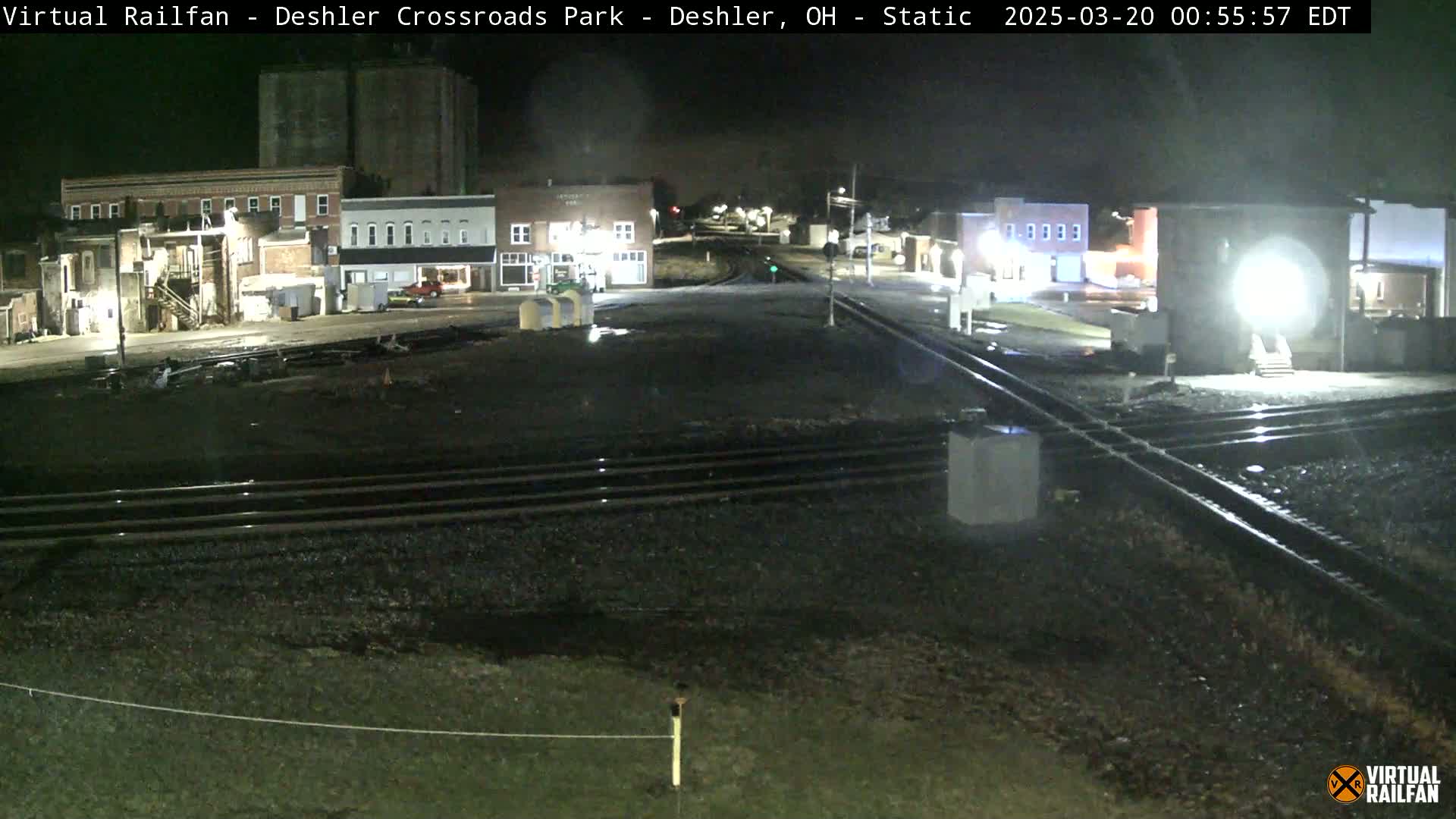 A nighttime, low-angle view of a railroad crossing in a small town, with several buildings visible in the background under dark conditions.