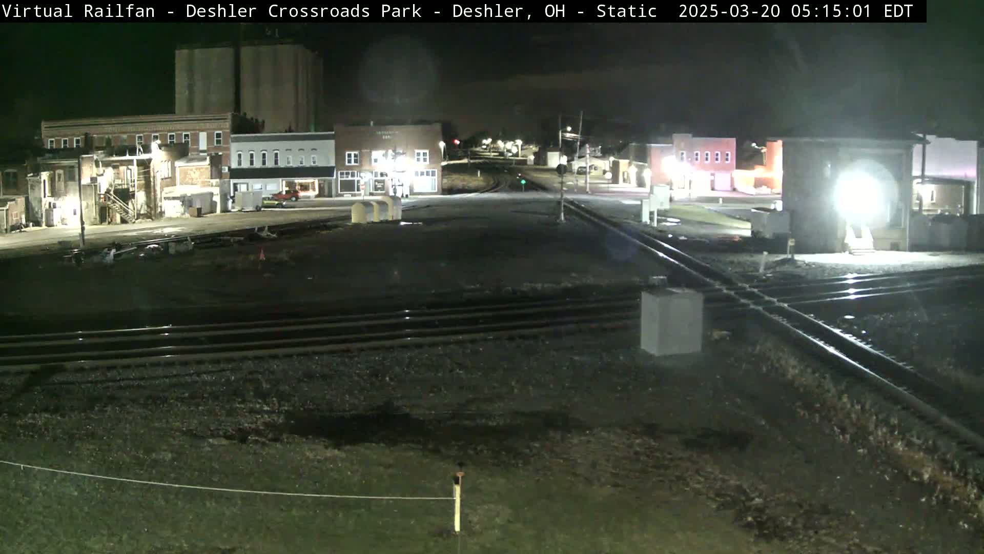 A nighttime view of railroad tracks intersecting a road in front of a small town under clear skies.