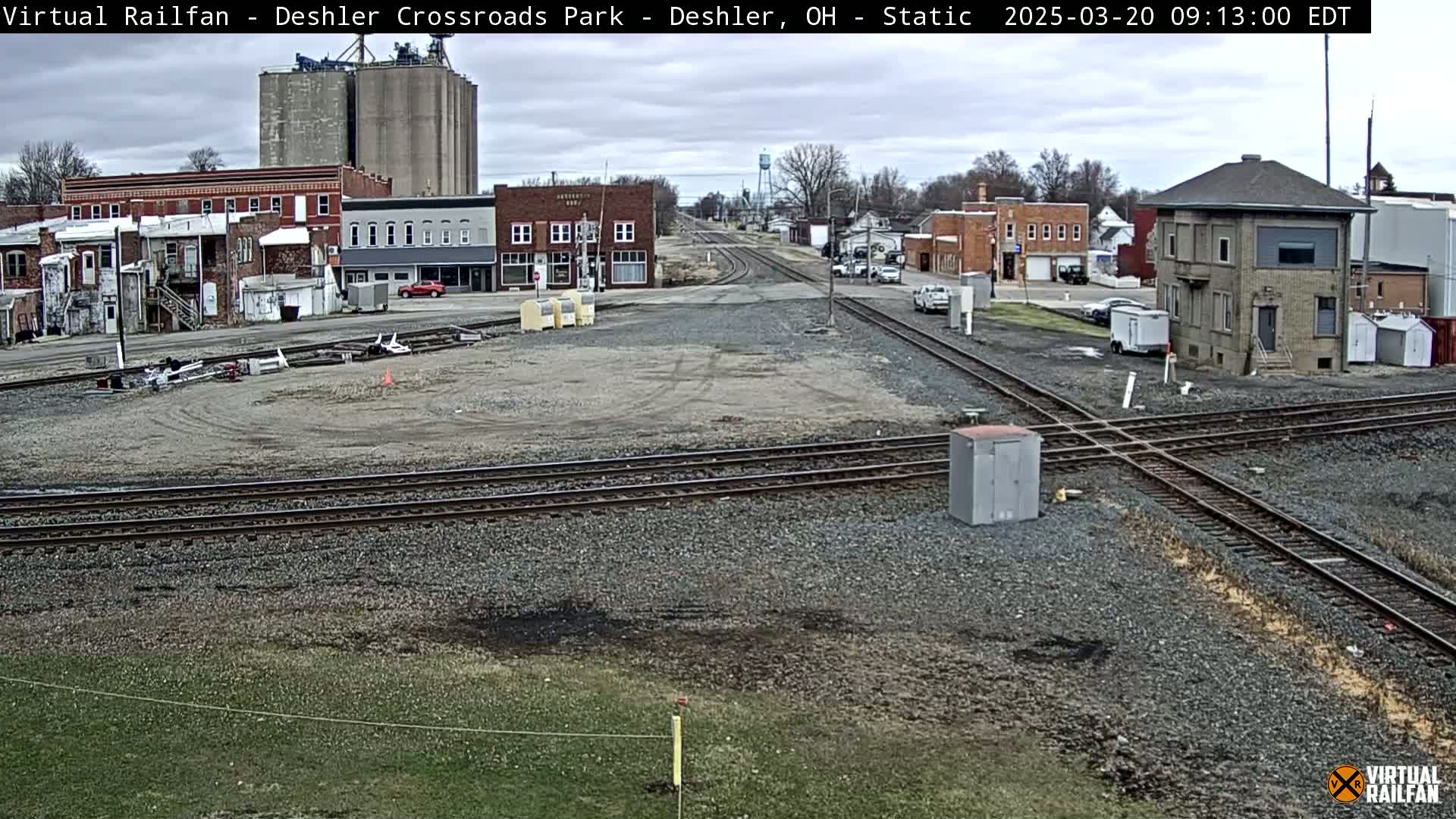 A railroad crossing in a small town under an overcast sky, with several buildings and grain silos in the background.