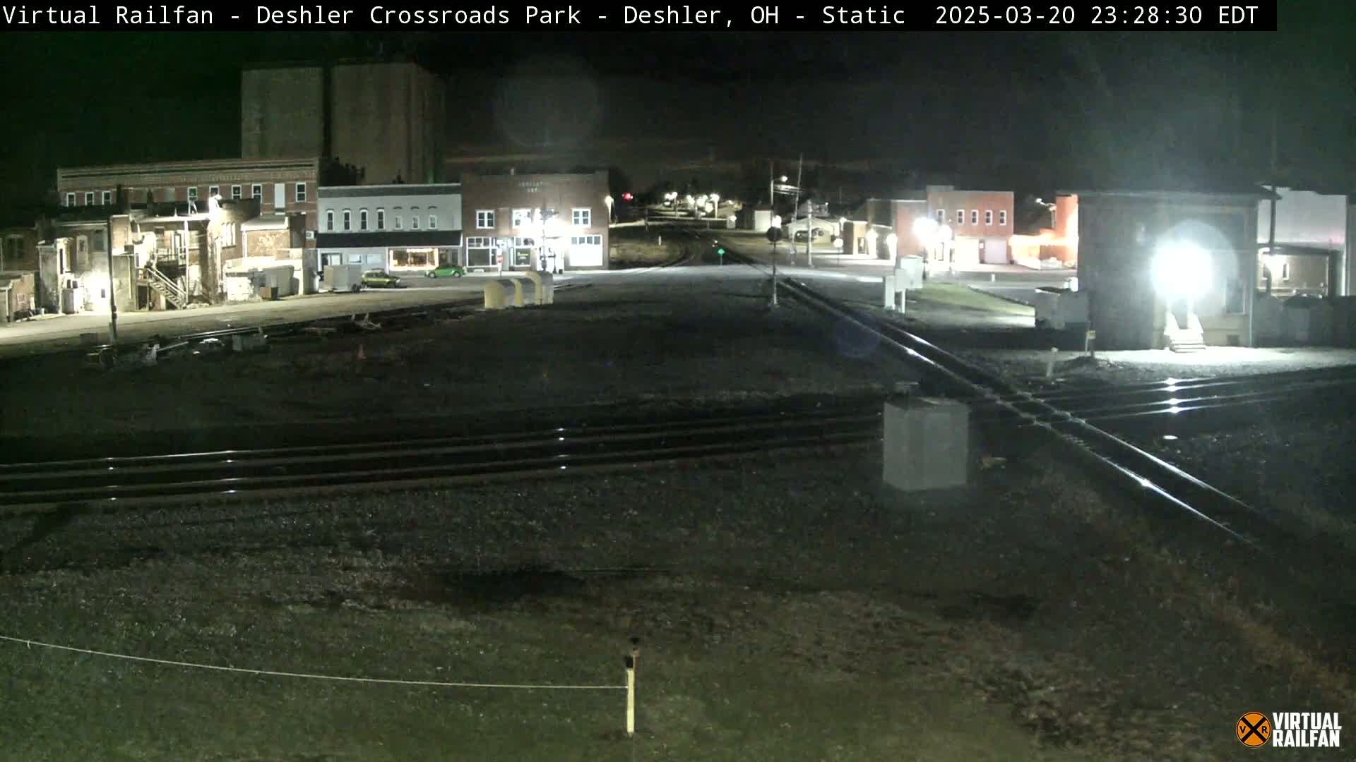 At night, a railroad crossing in a small town is shown, with several buildings and streetlights visible.