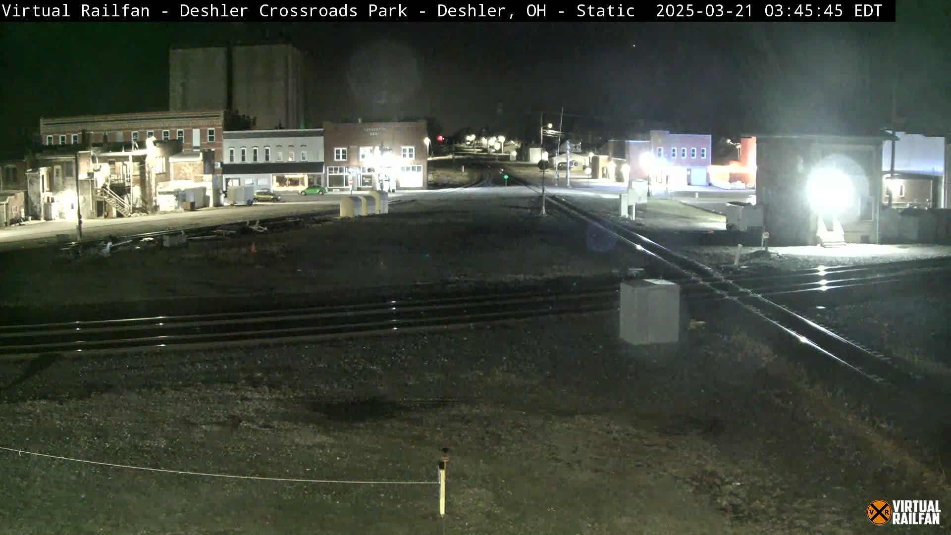A nighttime view of a railroad crossing in a small town, with several buildings illuminated under dark skies.