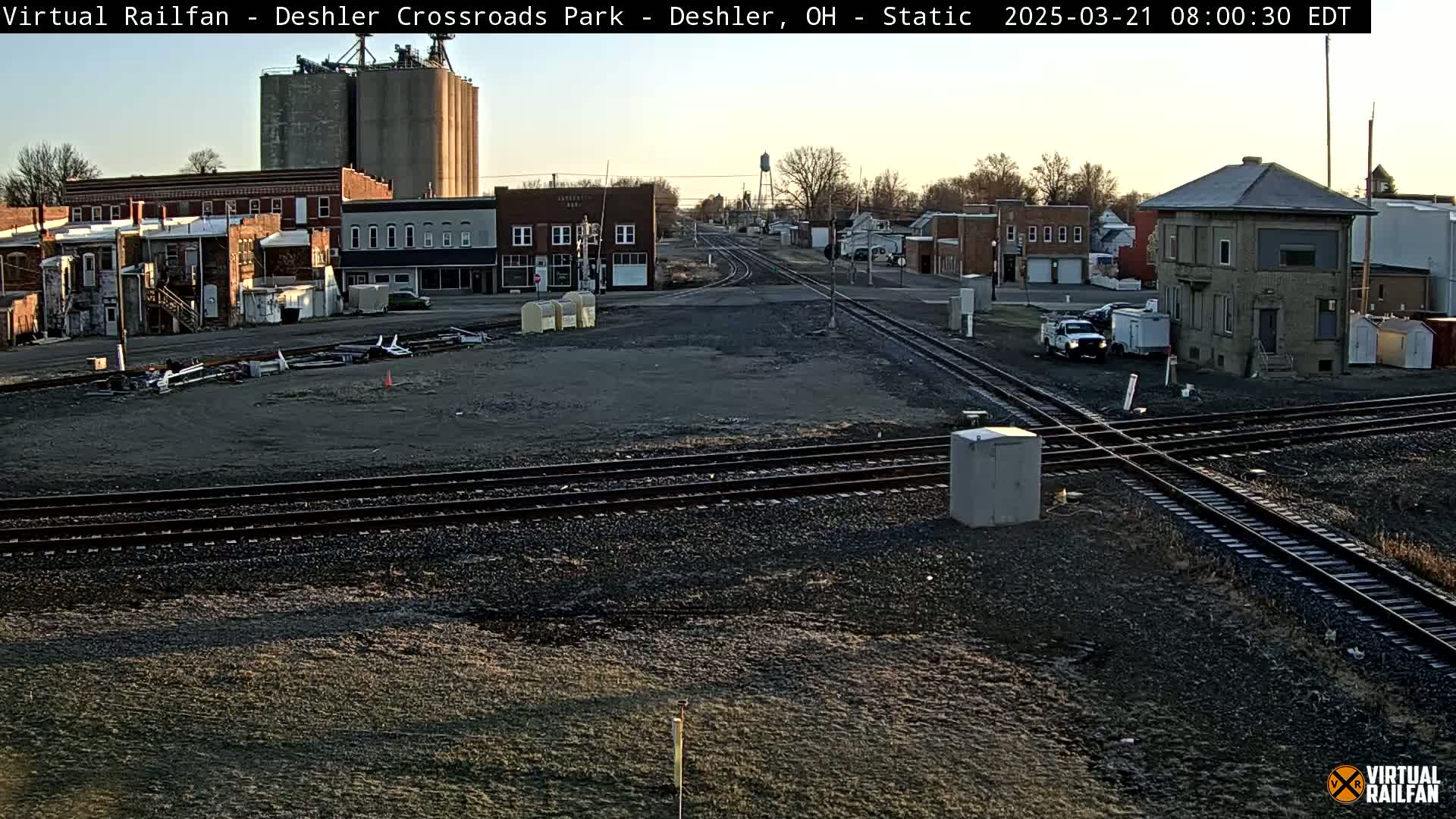 A train crossing in a small town under a mostly clear sky, with several buildings and grain silos in the background.
