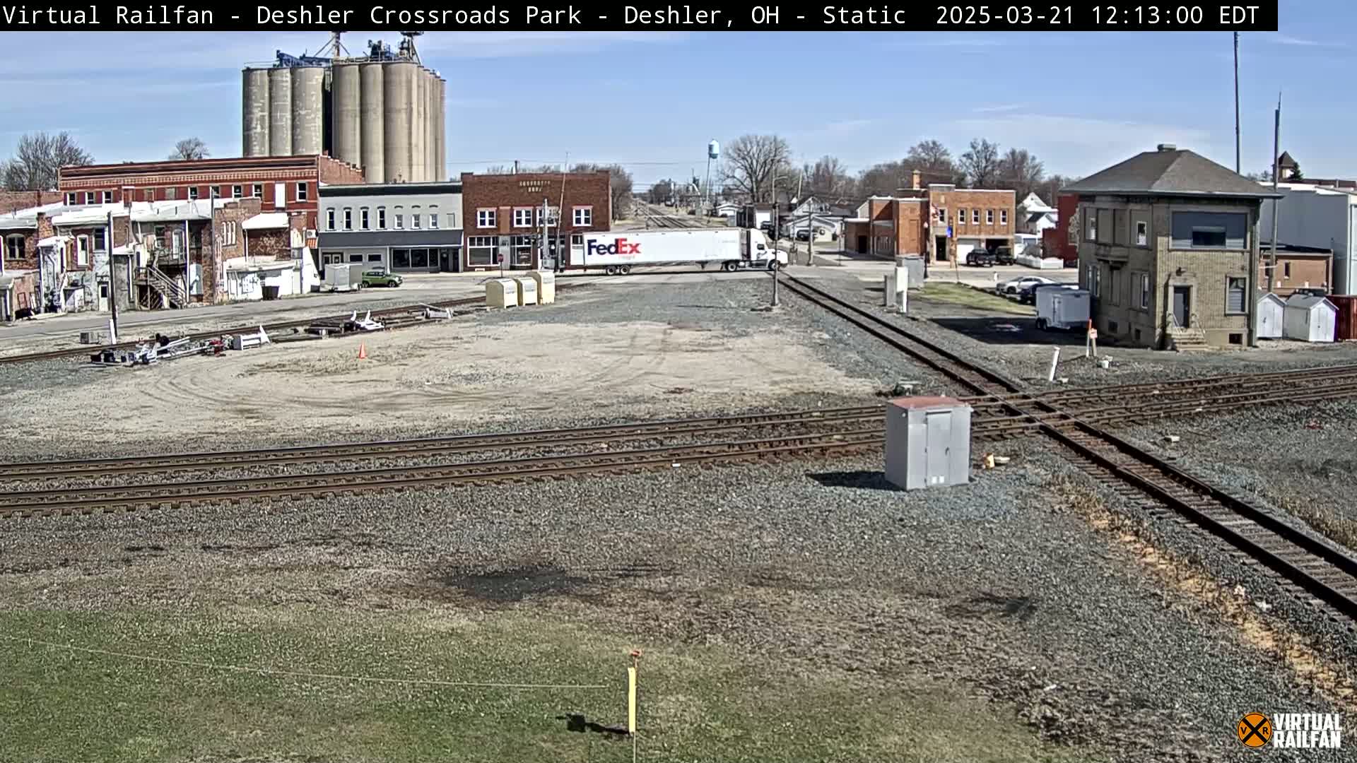 A sunny day reveals a railroad crossing in a small town, with a FedEx truck and grain silos visible in the background.