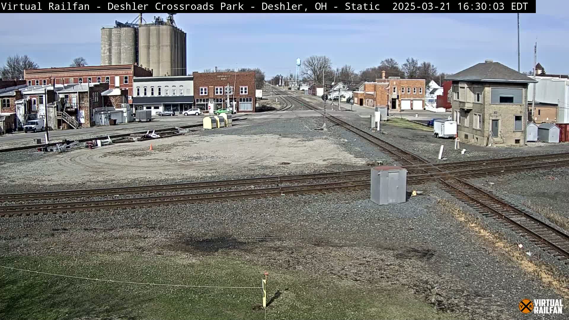 A train crossing in a small town on a sunny day, with railroad tracks intersecting in front of buildings including grain silos.