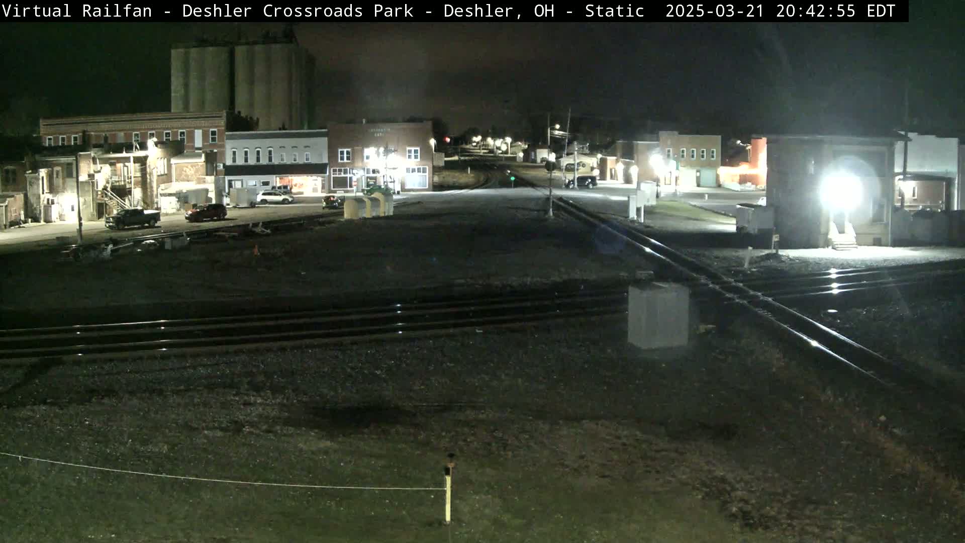 A nighttime view of a railroad crossing in a small town, with several buildings and a few vehicles visible under dark skies.