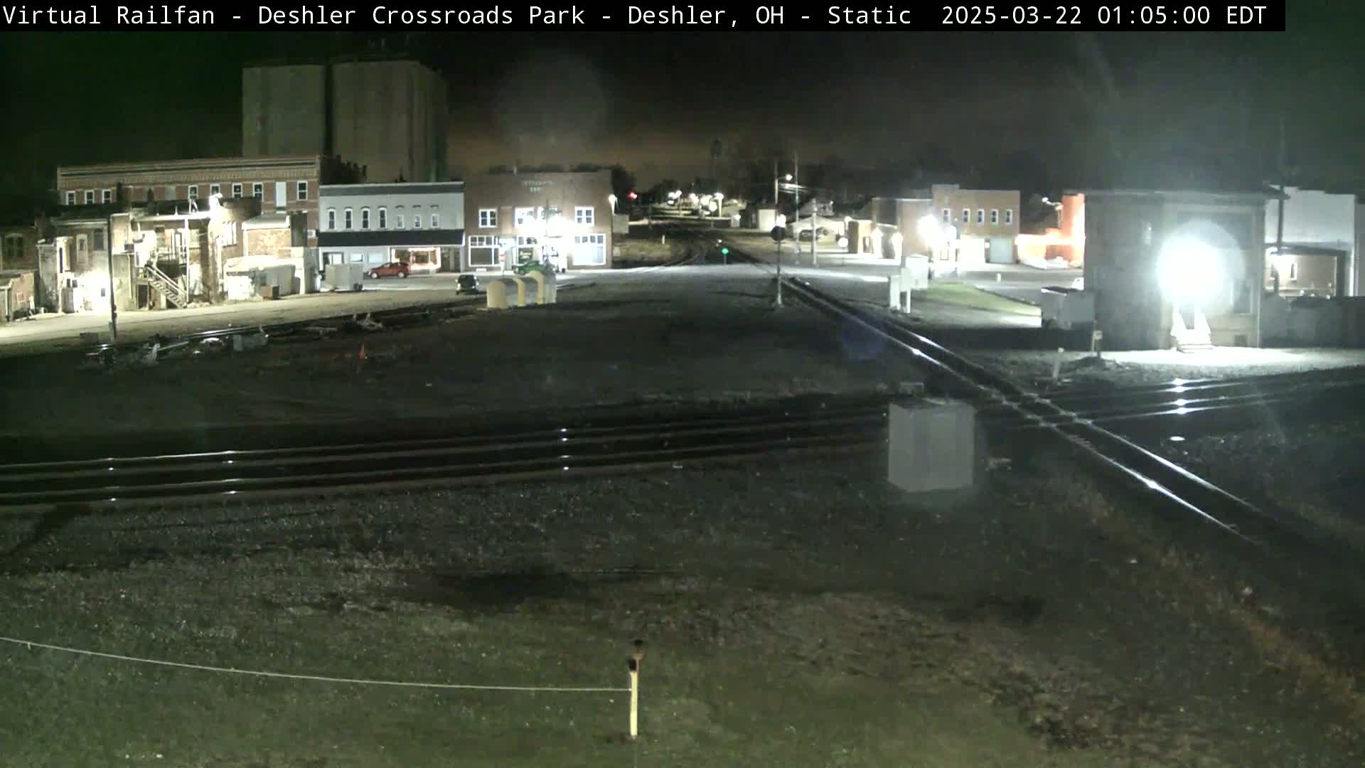 A nighttime view of a railroad crossing in a small town, with several buildings visible under a dark sky.