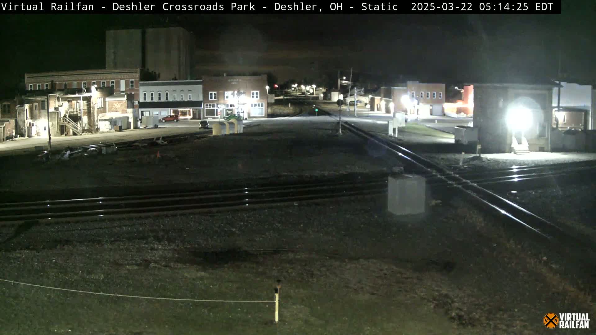 A nighttime view of a railroad crossing in a small town, with several brick buildings visible in the background under a clear sky.