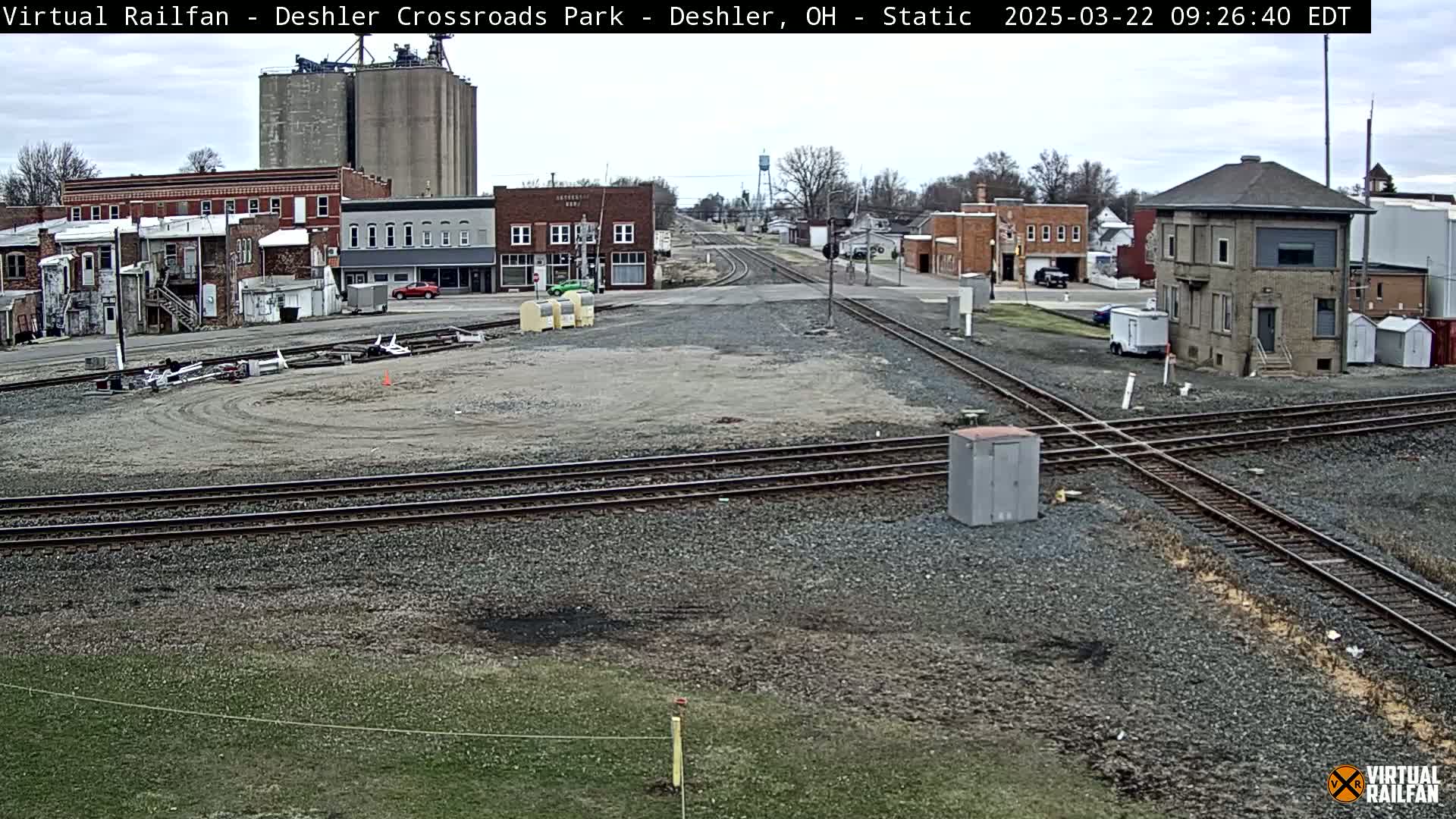 A mostly cloudy day shows a railroad crossing in a small town with several buildings, grain silos, and a train signal tower.