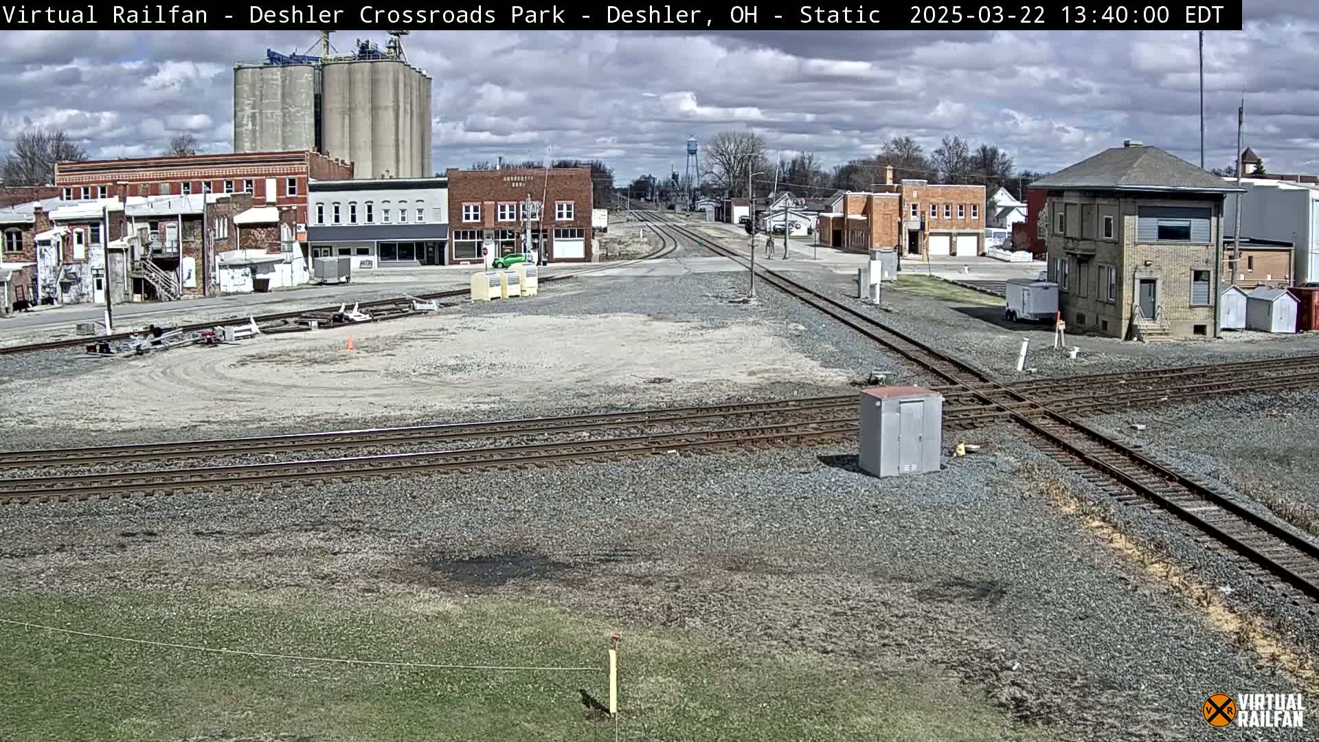A mostly clear day shows a train crossing in a small town with several buildings, grain silos, and train tracks intersecting.