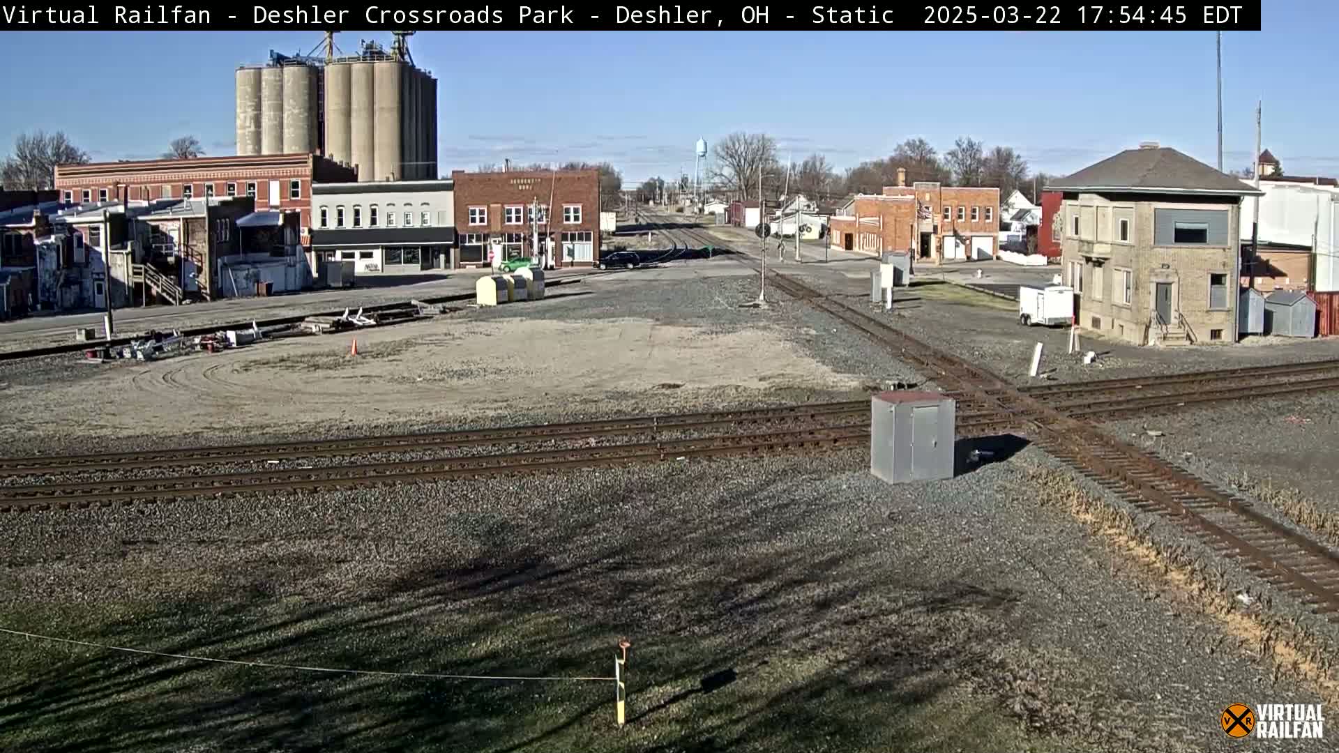 A sunny view of a railroad crossing in a small town with several buildings, grain silos, and a rail signal tower.