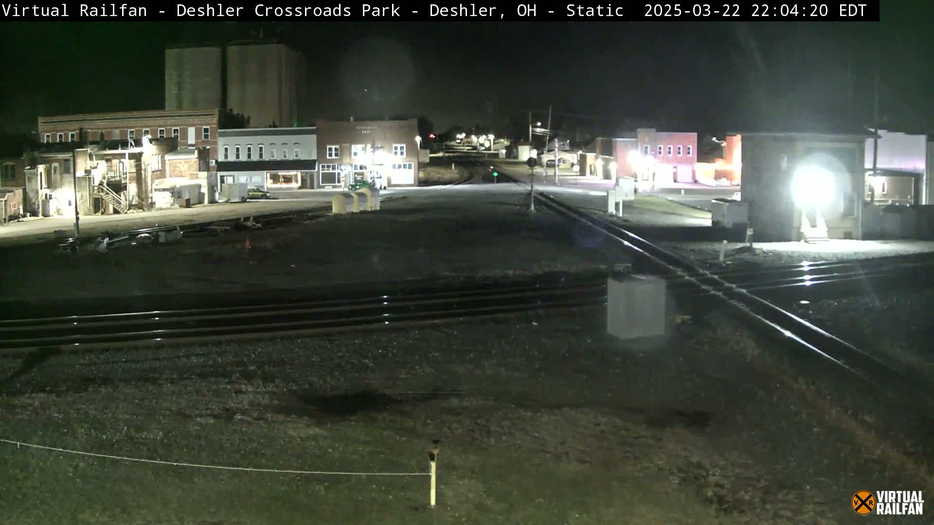A nighttime view of a railroad crossing in a small town, with buildings and grain silos visible in the background.