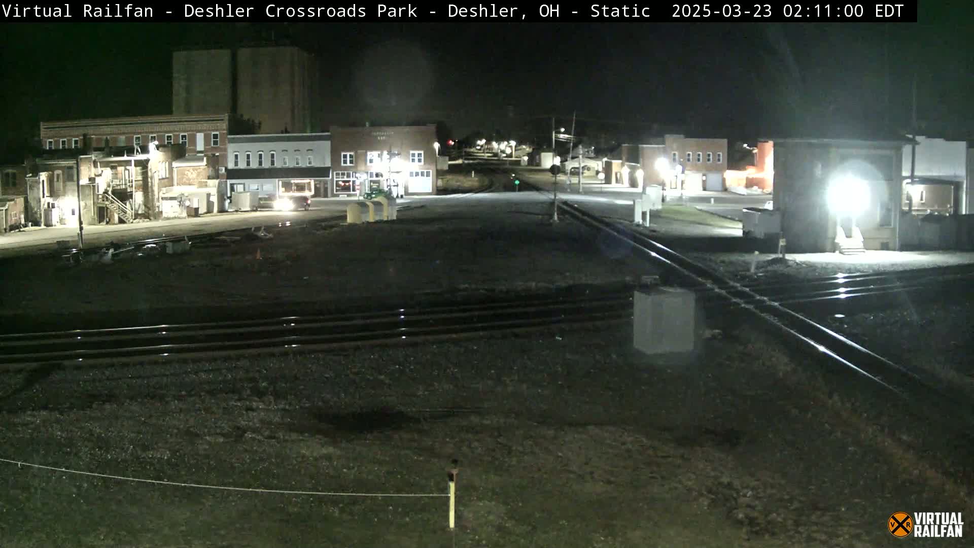 At night, a nighttime view shows a railroad crossing in a small town with several buildings and streetlights.