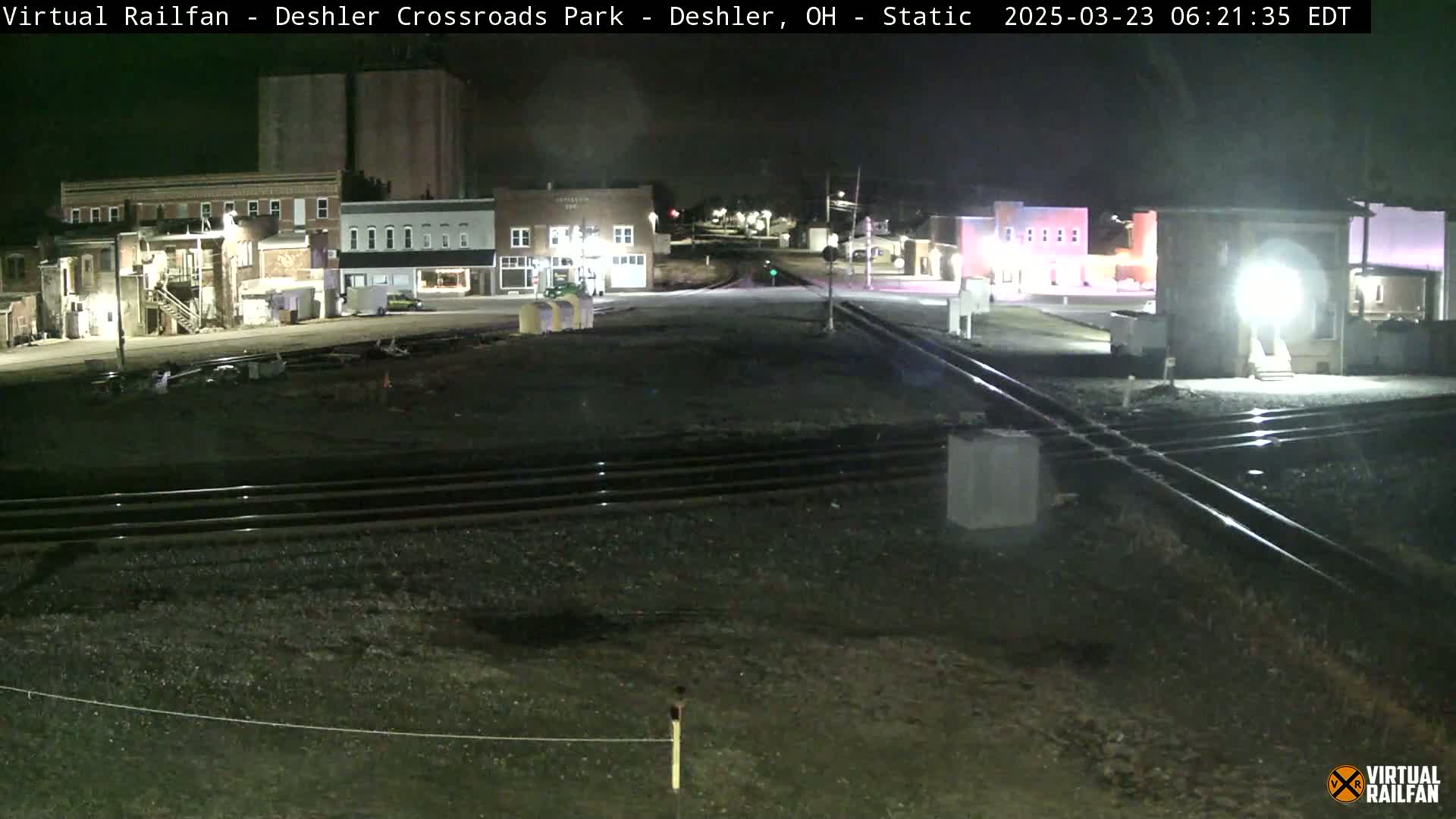 A nighttime view of a railroad crossing in a small town, with several buildings illuminated under the dark sky.