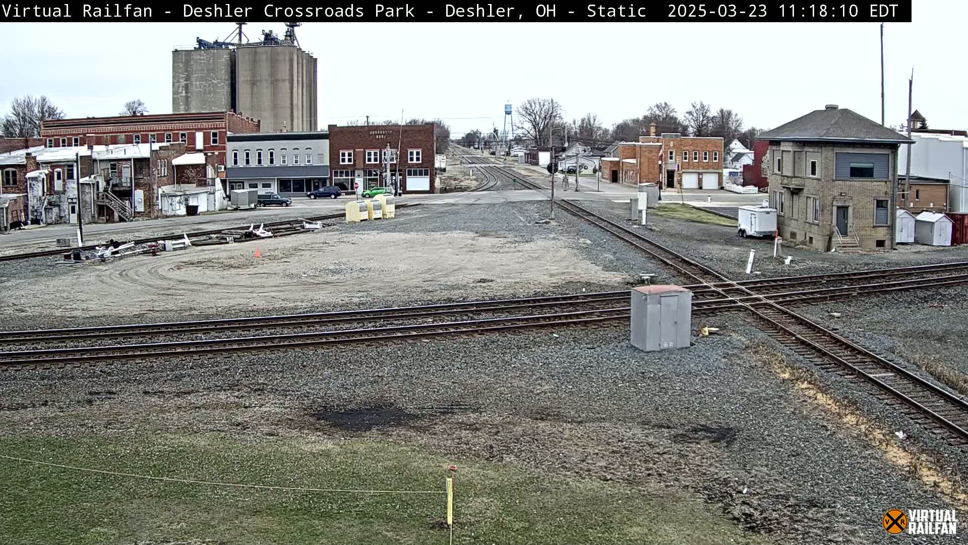 A railroad crossing in a small town on an overcast day, with several buildings and grain silos visible in the background.